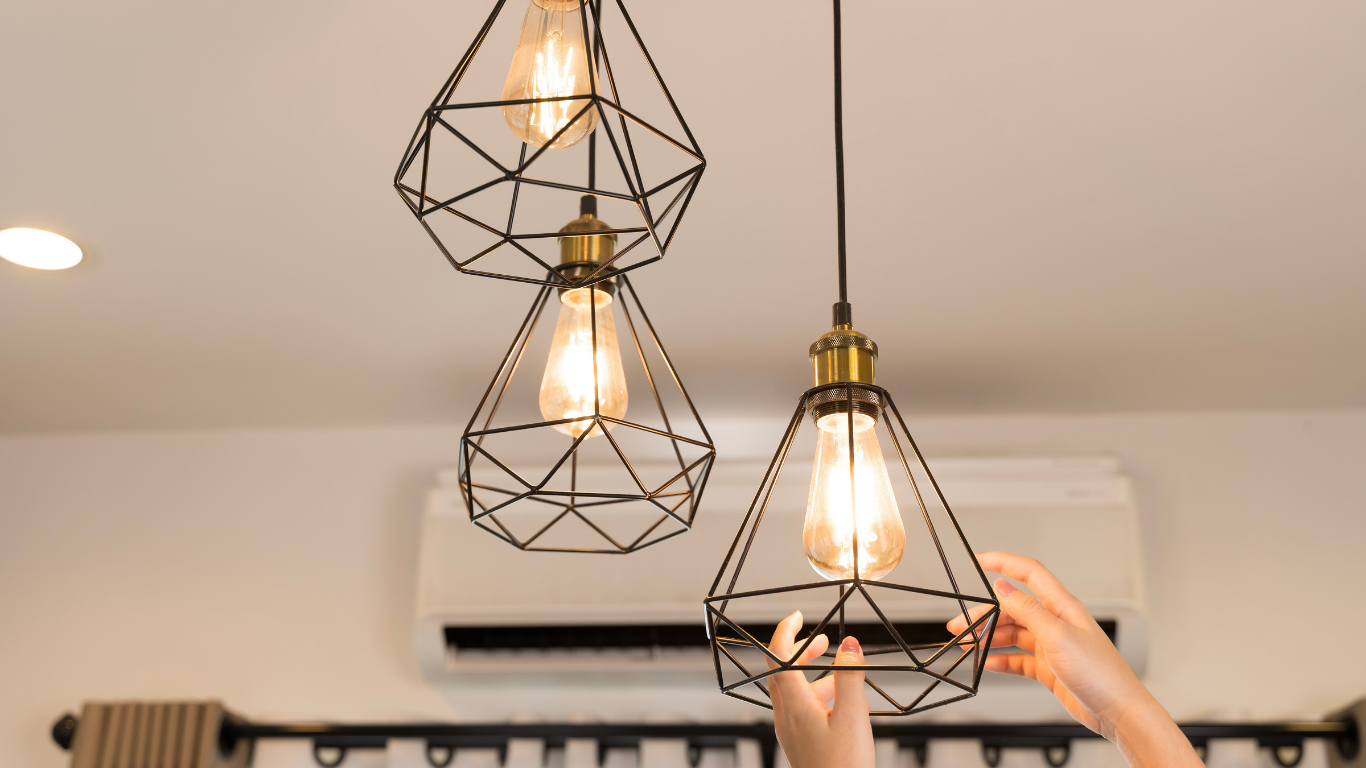 Three geometric pendant lights with warm bulbs hanging in a room, one being adjusted by a hand.