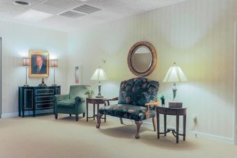 Small formal sitting room with chairs, lamps, a table, and a framed portrait on the wall.