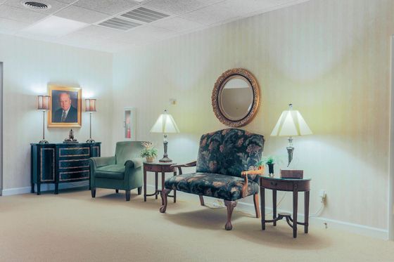 Small formal sitting room with chairs, lamps, a table, and a framed portrait on the wall.