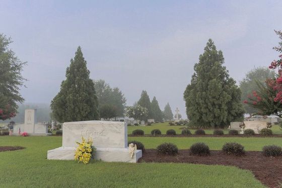 Graveyard with white headstones, green lawns, and tall trees under a hazy sky.