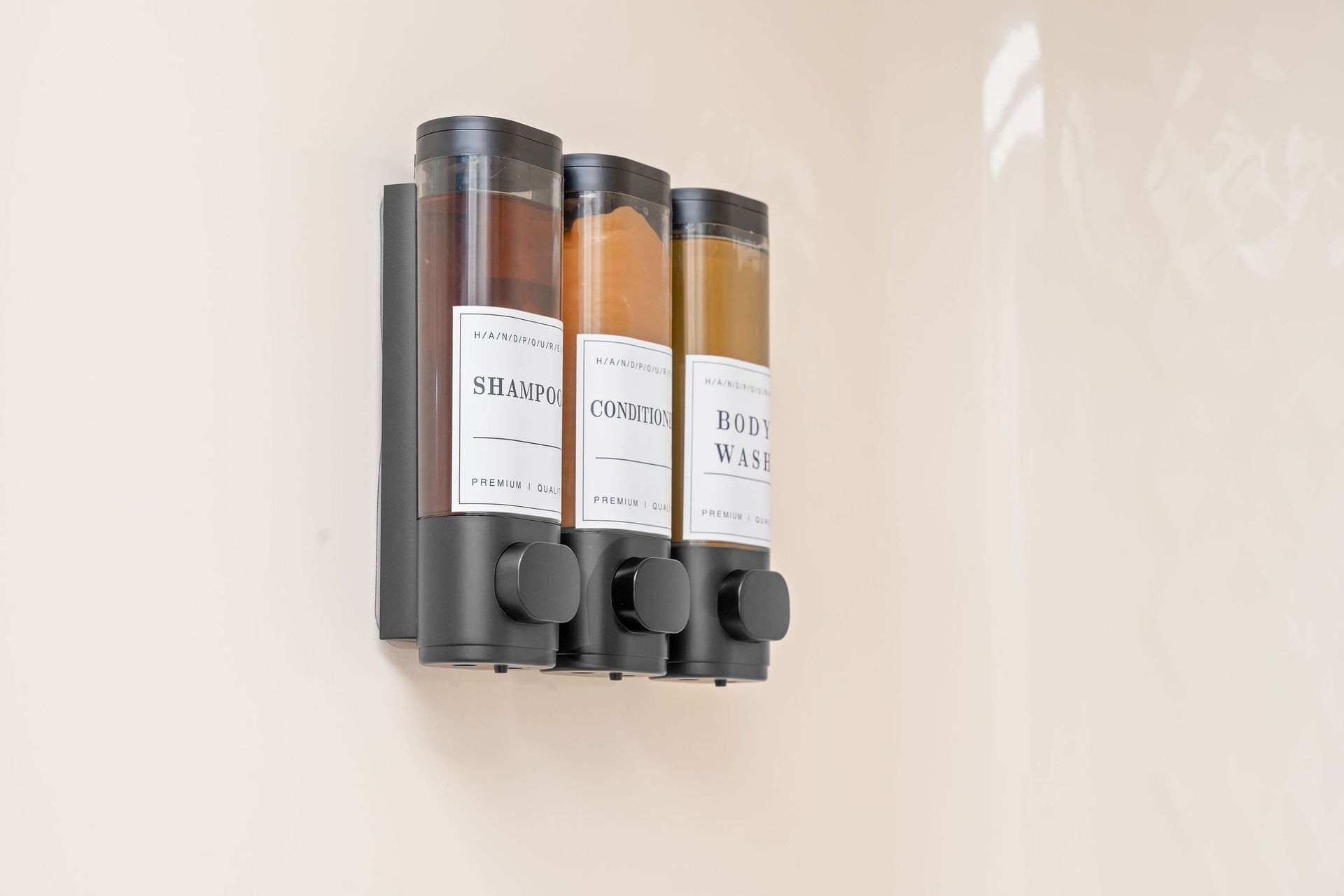 A wall-mounted triple soap dispenser unit with labels, mounted on a plain, light-colored shower wall.