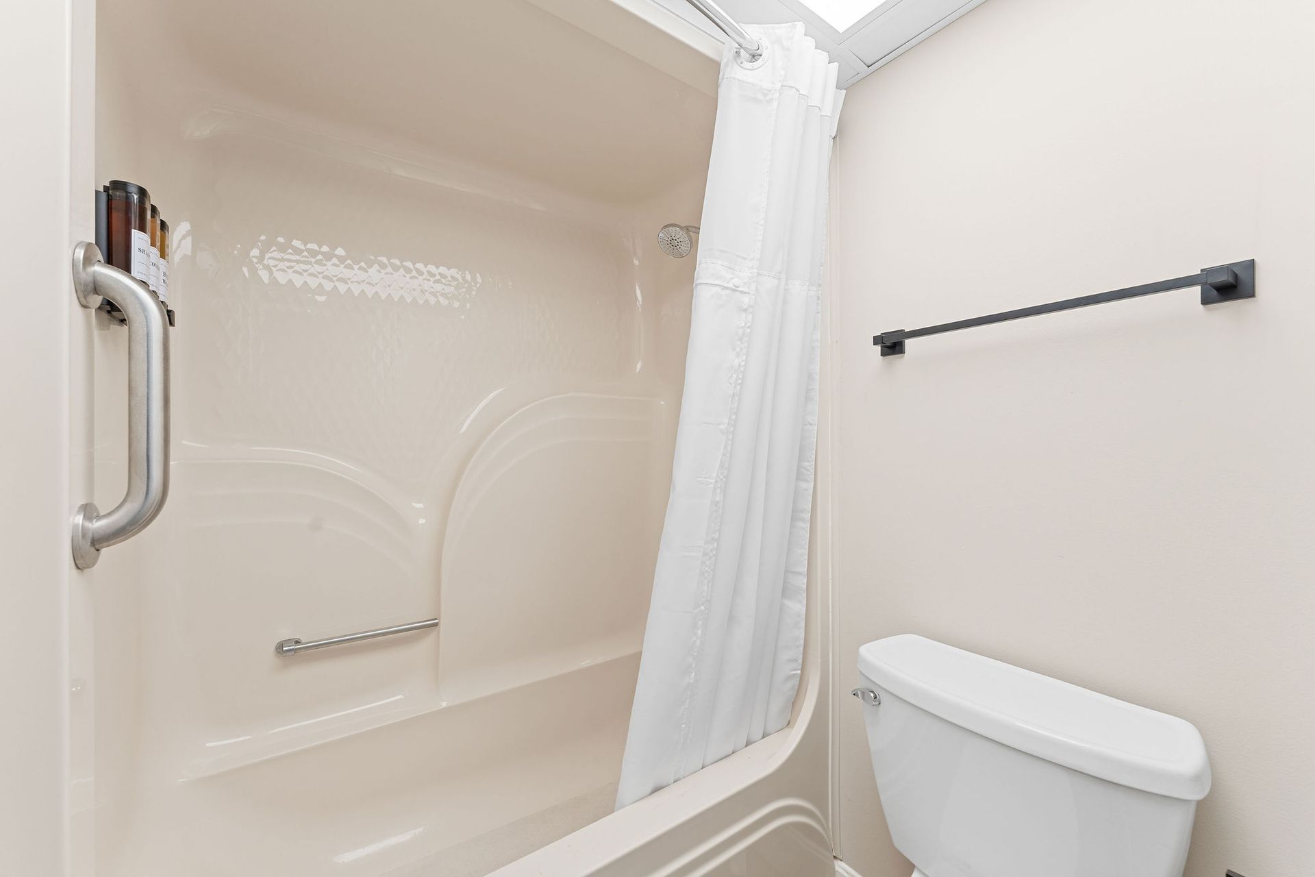 A light beige shower with a grab bar and white curtain next to a white toilet with a black towel bar on the wall.