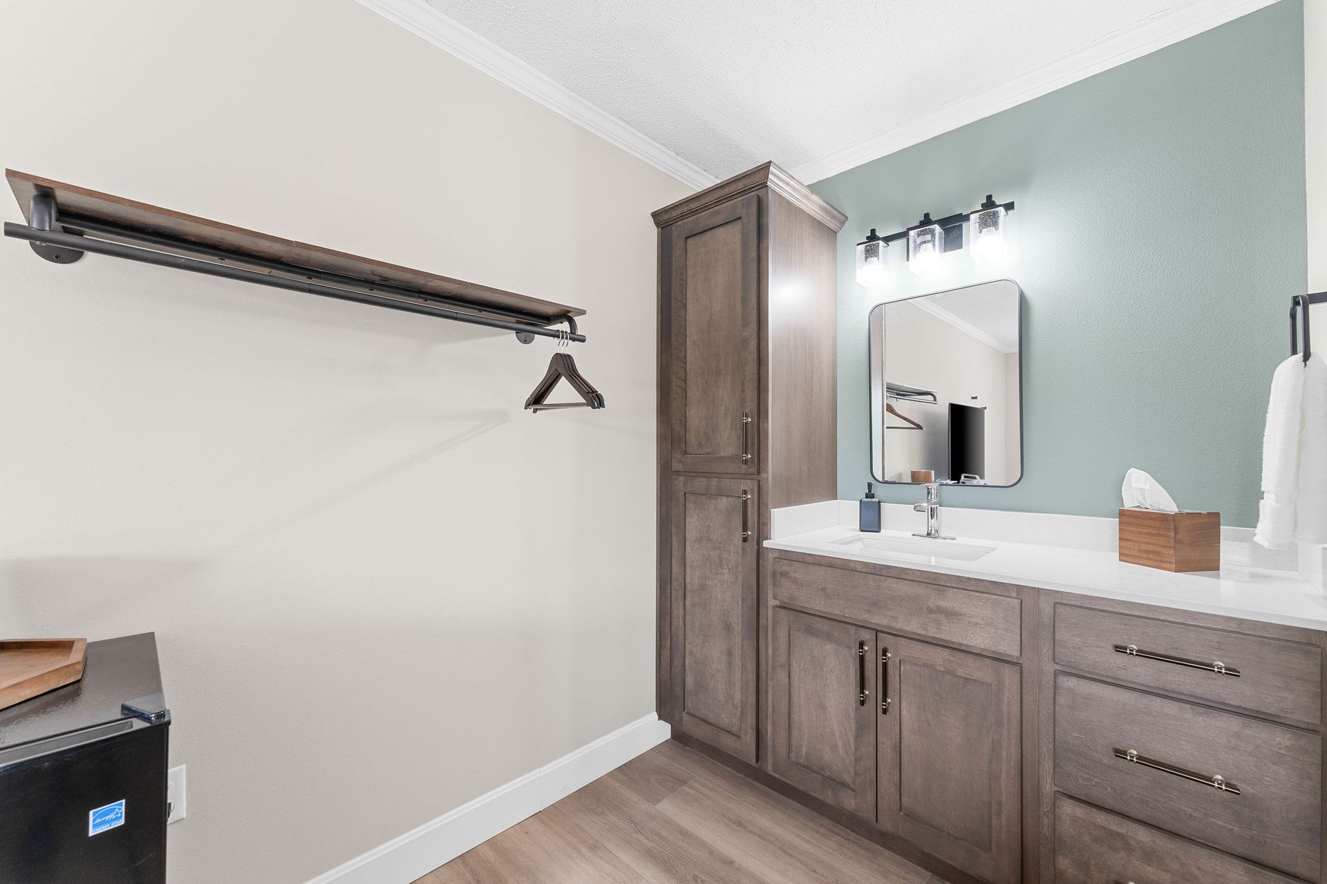 A bathroom vanity with dark wood cabinets, a teal accent wall, a mirror, and a wall-mounted clothing rack to the left.