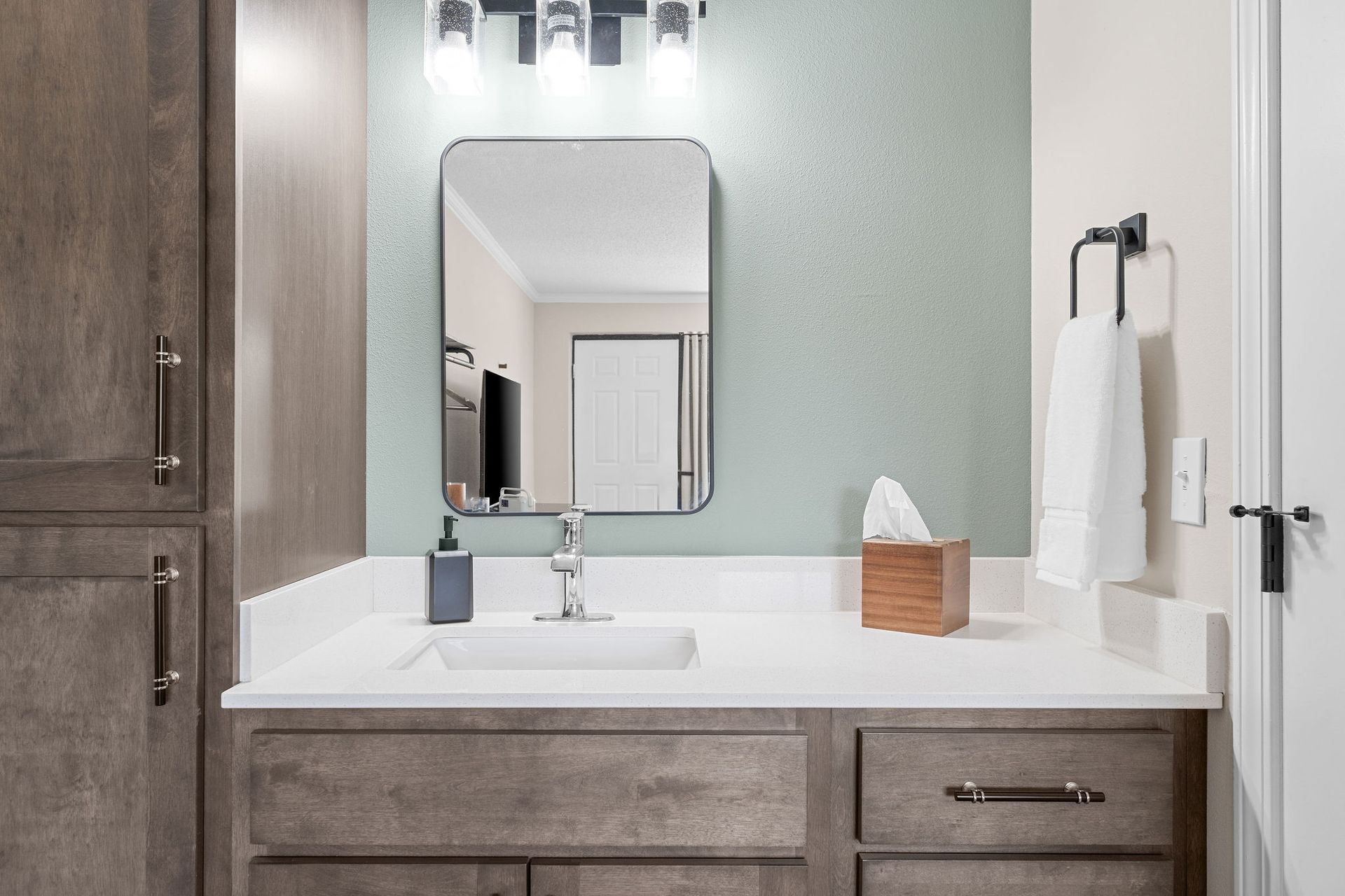 Modern bathroom vanity with a wood cabinet, white countertop, rectangular mirror, and sage green wall.