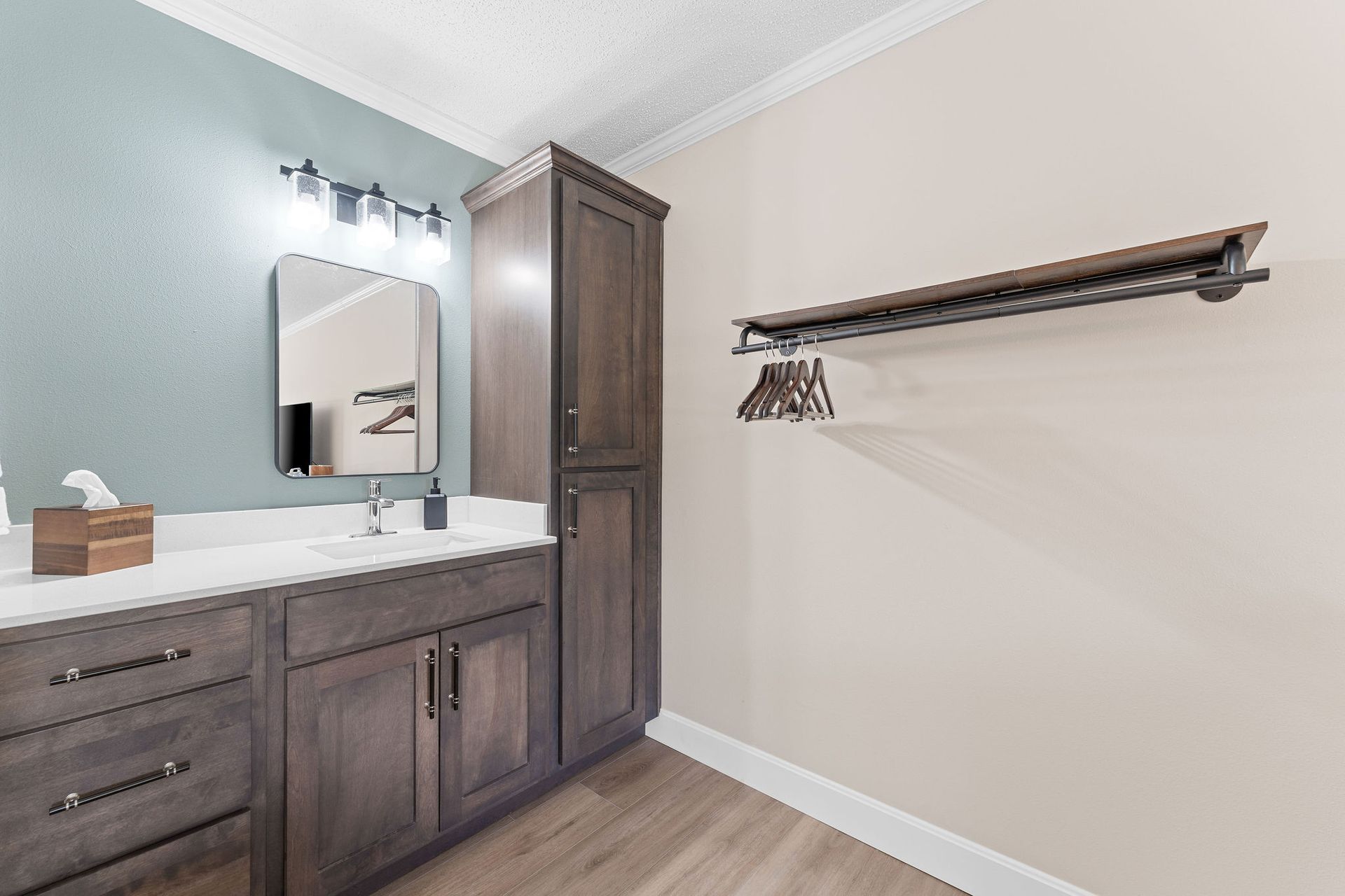 A vanity with a dark wood cabinet, white countertop, and mirror next to a wall-mounted clothing rack in a bathroom.