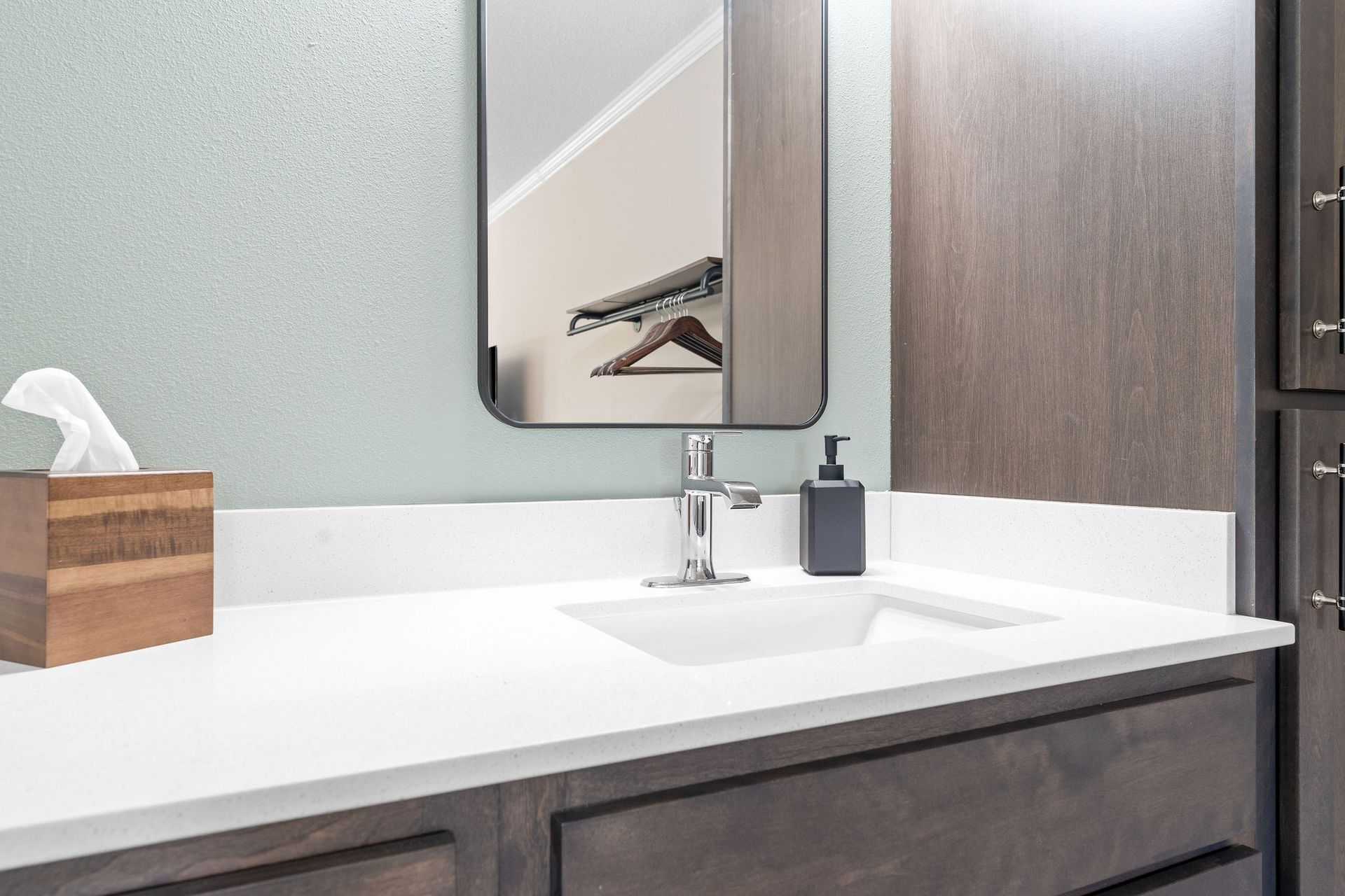 A modern bathroom vanity with a white countertop, chrome faucet, grey soap dispenser, and wooden tissue box.