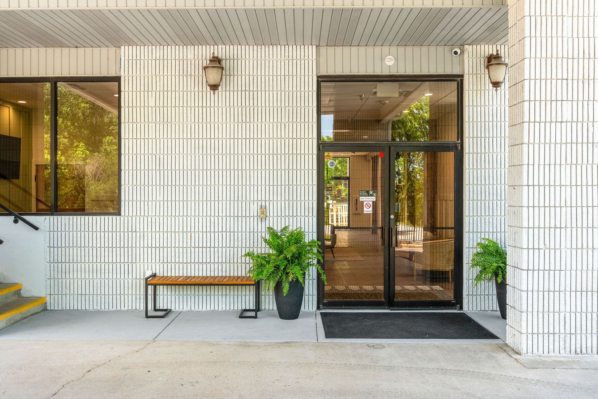 An entrance with white patterned brick walls, a wooden bench, a potted plant, and double glass doors under an overhang.