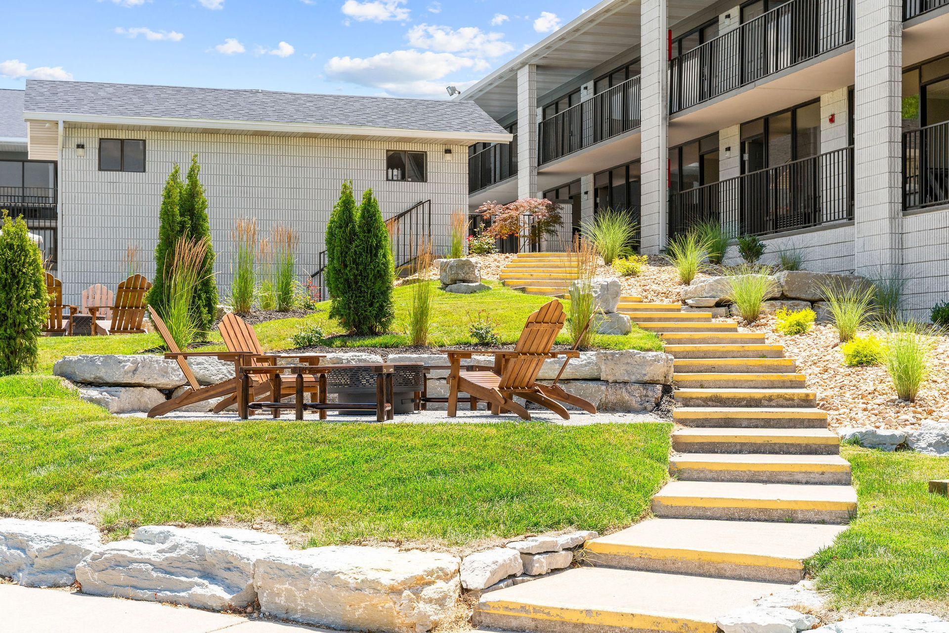 A terraced patio area with wooden chairs, a fire pit, and stone stairs leading to a multi-story building under a blue sky.