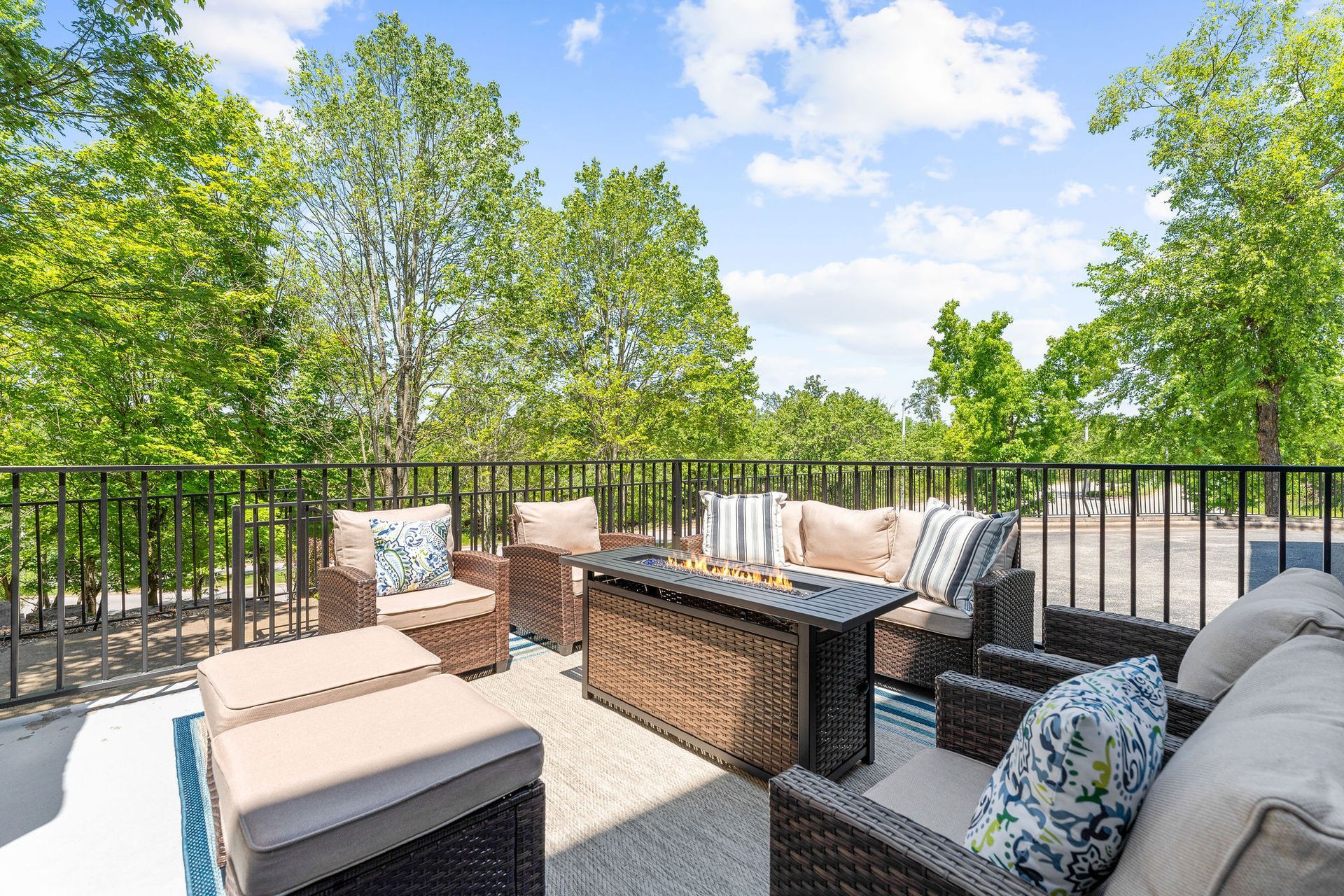 Outdoor patio with wicker furniture, a fire pit table, and patterned cushions, overlooking a tree-filled landscape.