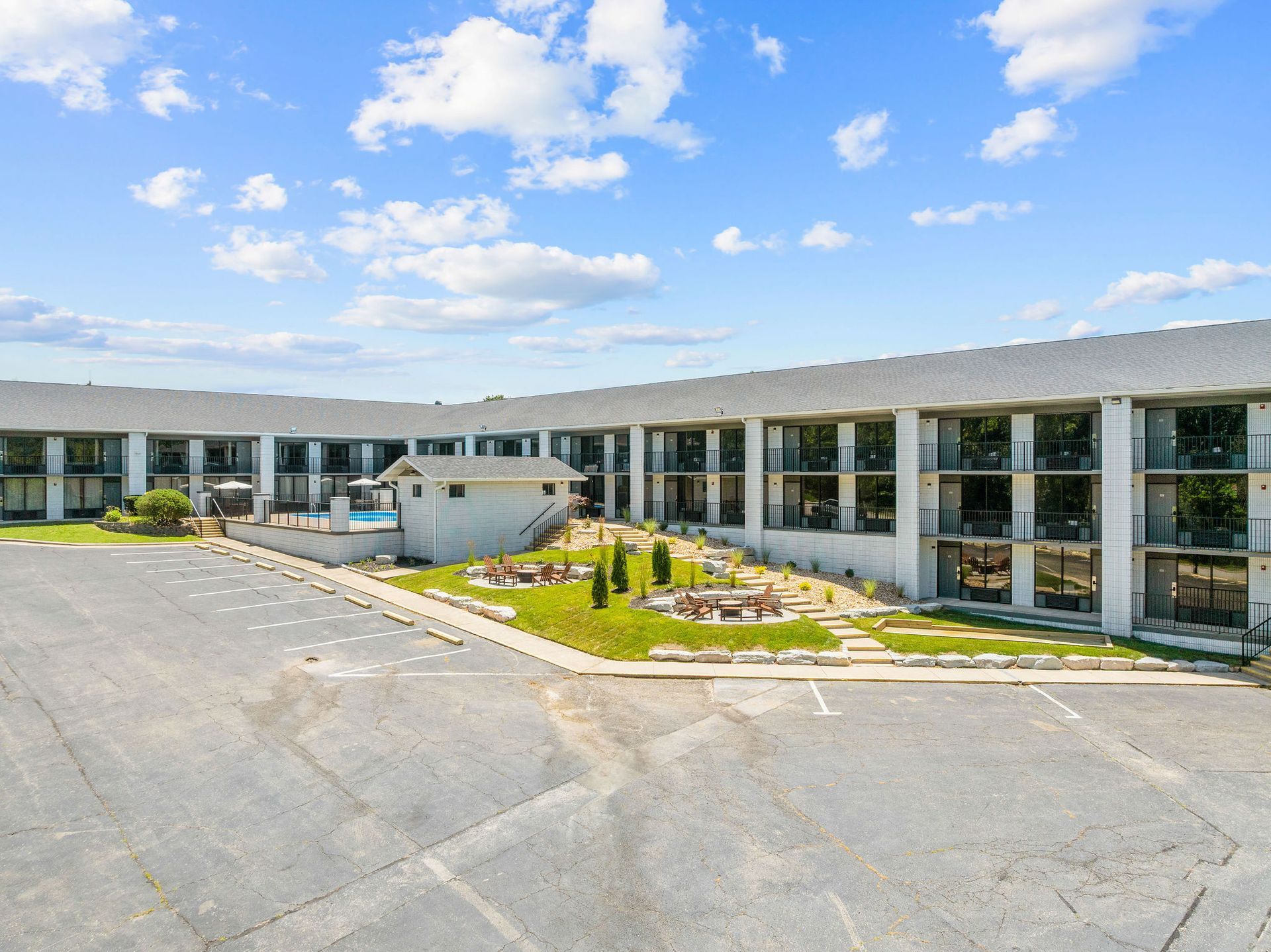 A wide exterior view of a two-story motel with a parking lot, a grassy courtyard, and a small pool under a blue sky.