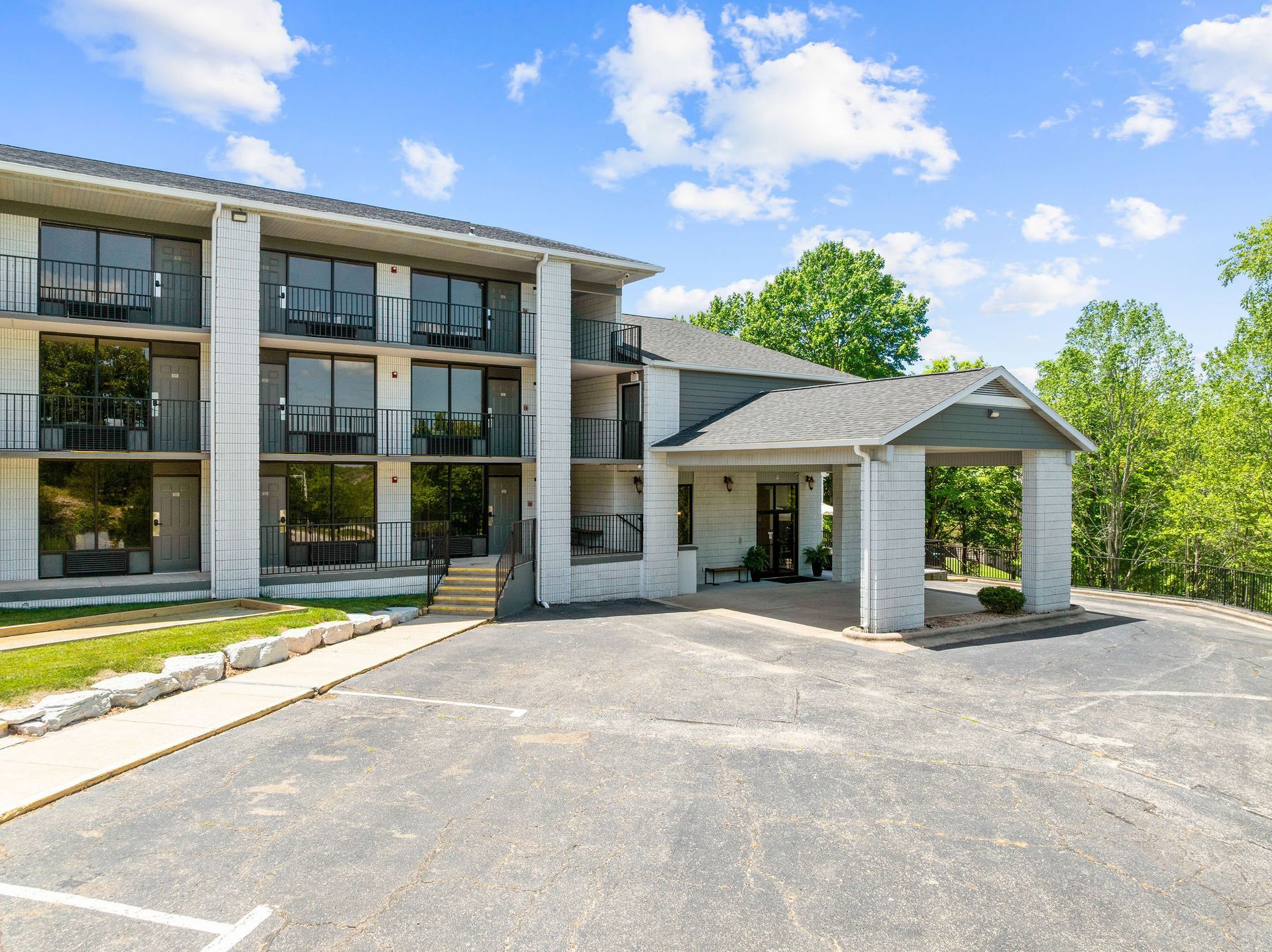 A three-story white brick building with a gravel parking lot under a sunny blue sky with trees in the background.
