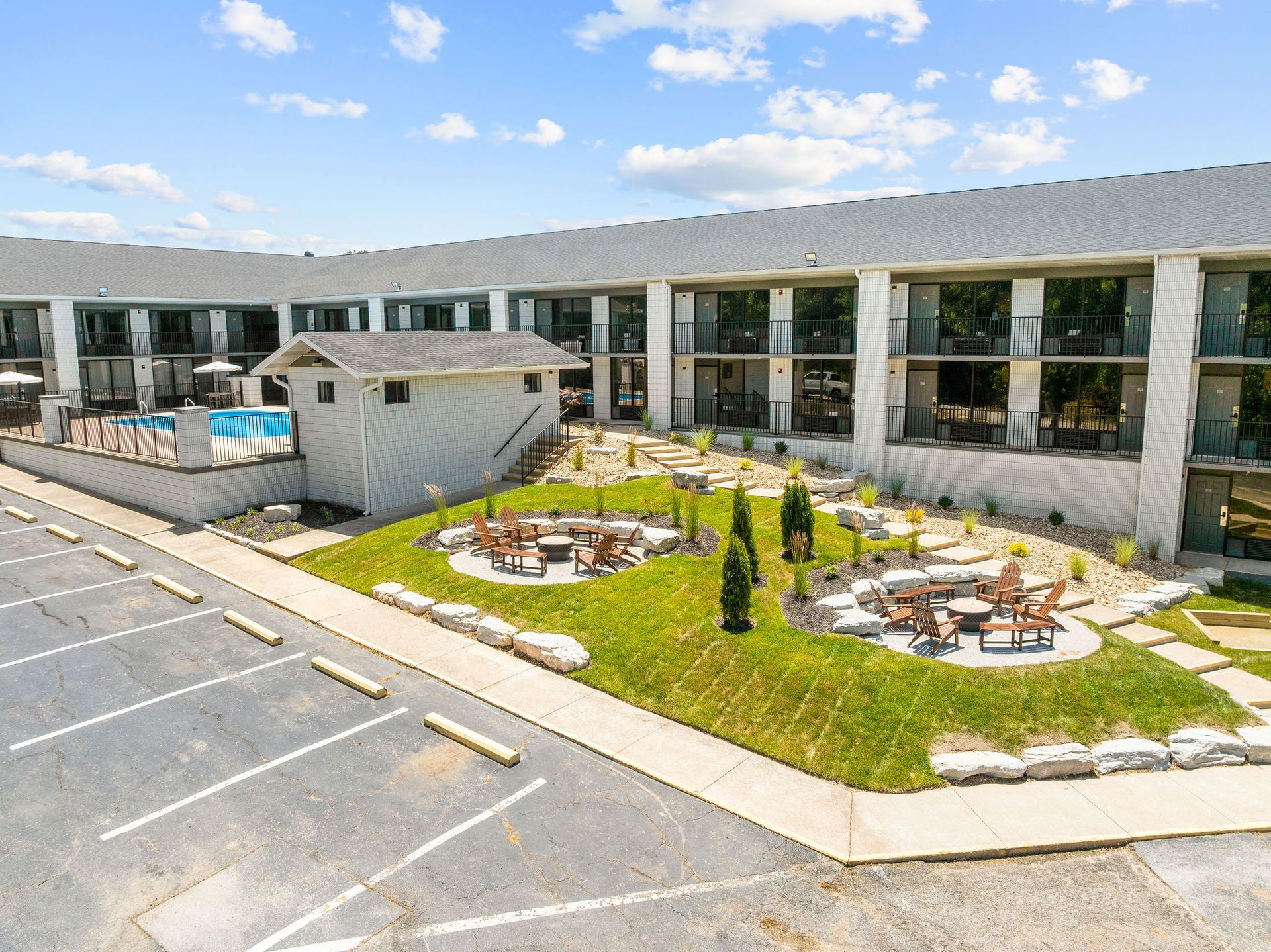 A modern motel building with an outdoor pool, a small stone shed, and two seating areas with fire pits on a grassy lawn.