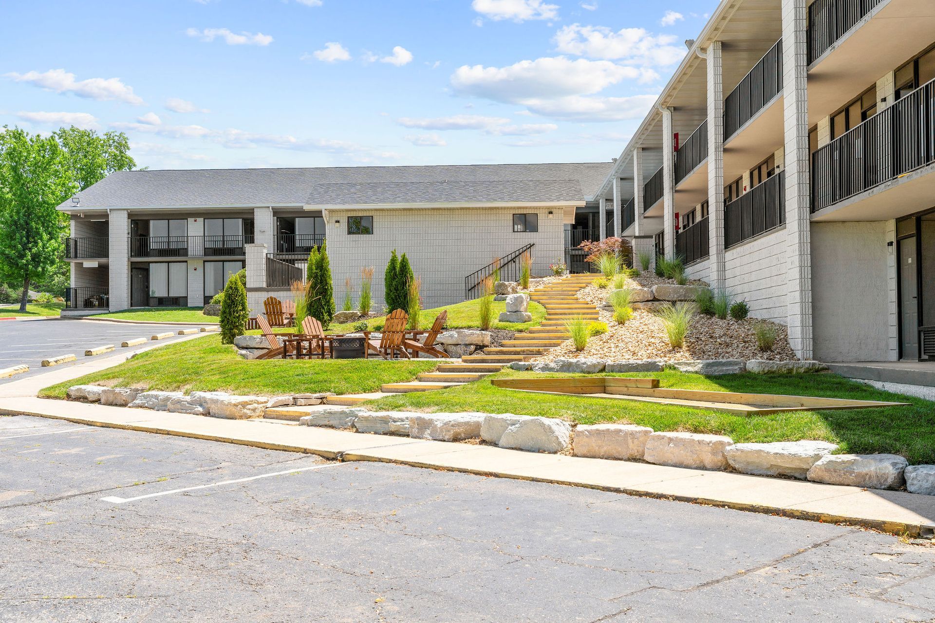 A multi-story hotel with a grey exterior, featuring a stone-retaining wall and a grassy courtyard with wooden chairs.