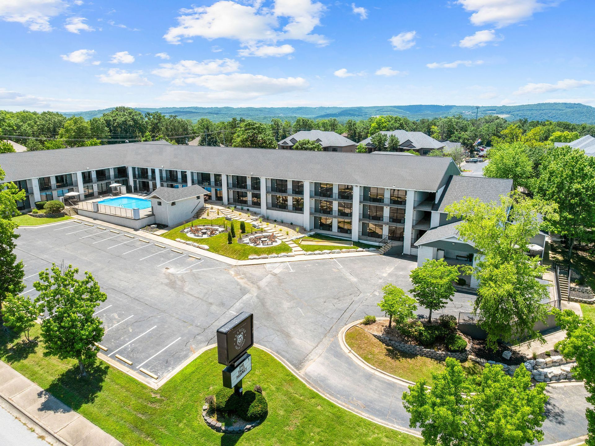 An aerial view of a multi-story, light-colored motel with a pool, parking lot, and surrounding trees under a blue sky.