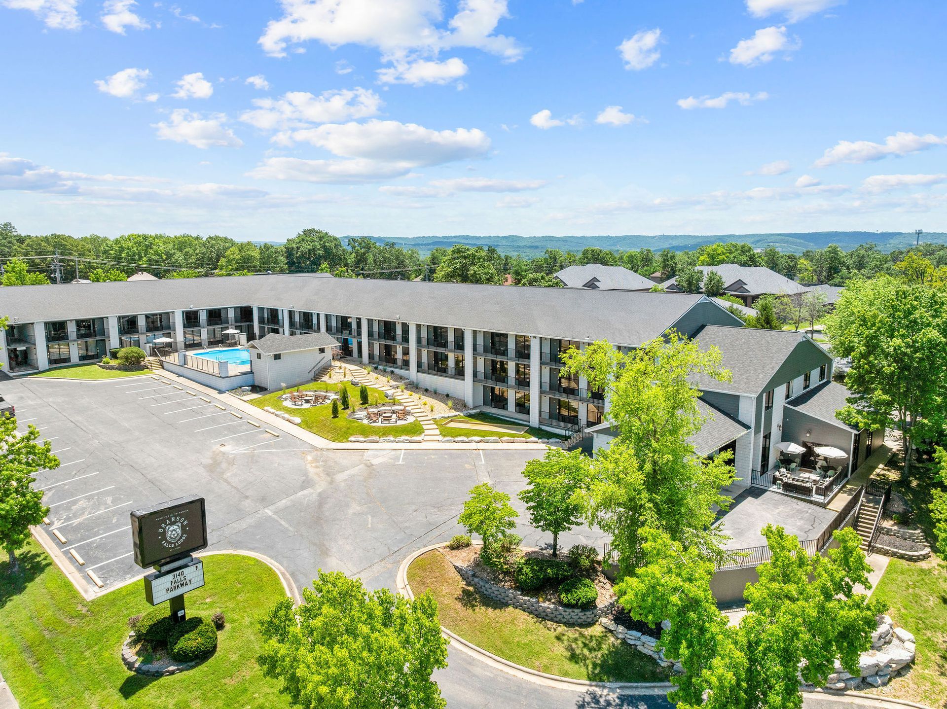 An aerial view of a multi-story hotel with a parking lot and outdoor swimming pool surrounded by green trees.