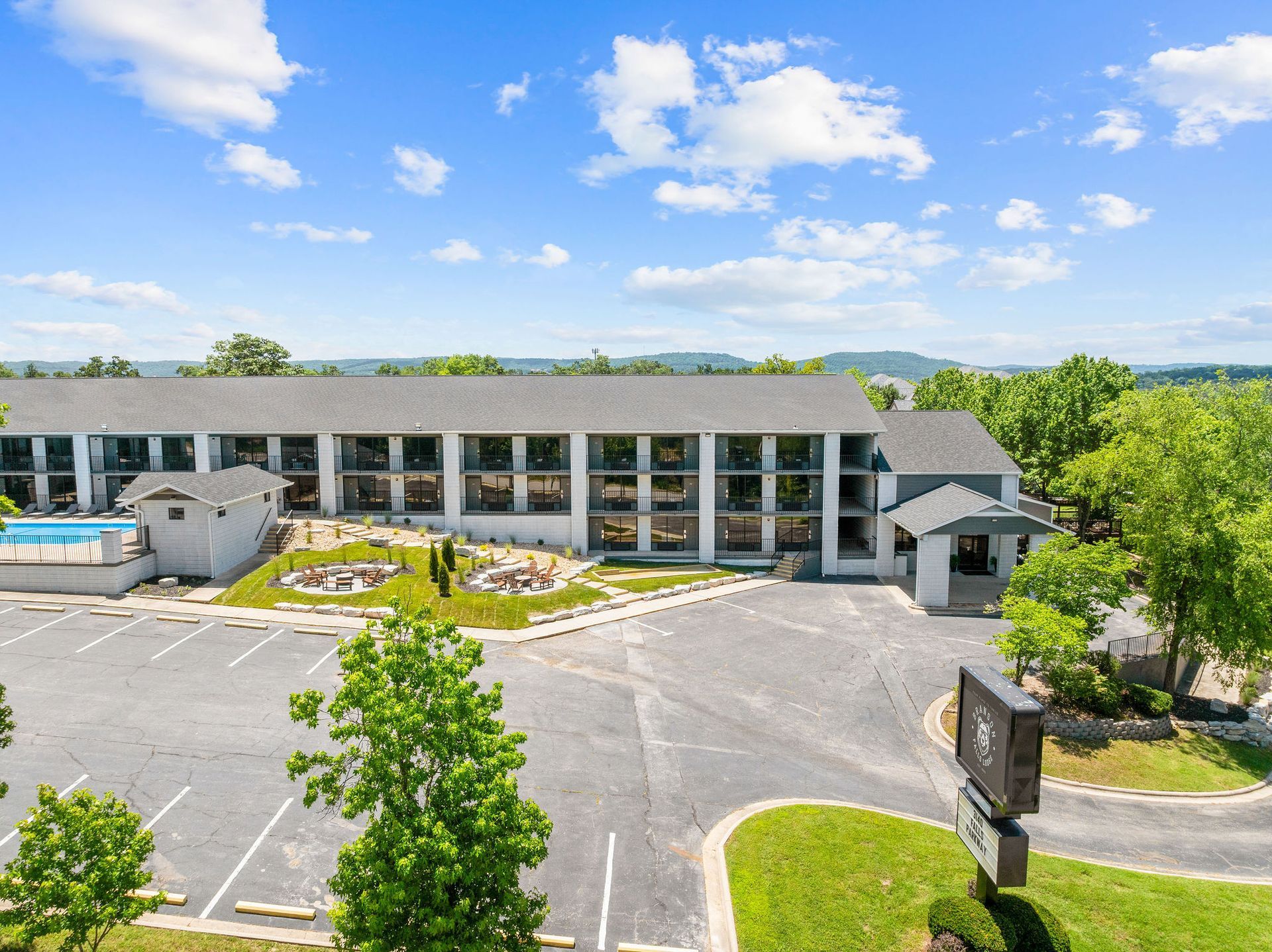 An elevated view of a multi-story hotel building with a parking lot, swimming pool, and trees under a sunny blue sky.