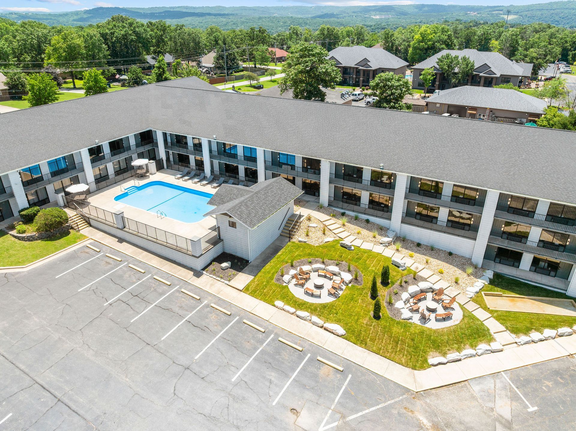 Aerial view of a motel with a swimming pool, parking lot, and outdoor seating area surrounded by trees and residential homes.