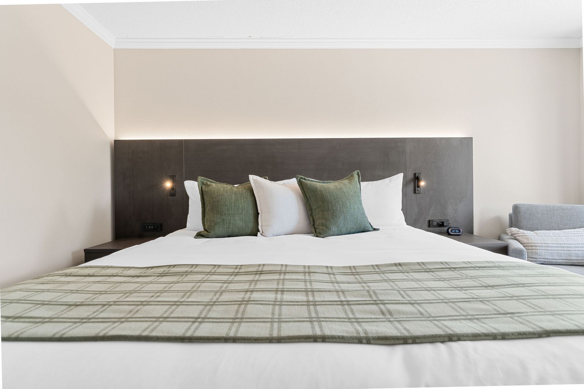 A bed with a plaid runner and sage green pillows in a modern bedroom with a dark wooden headboard and light walls.