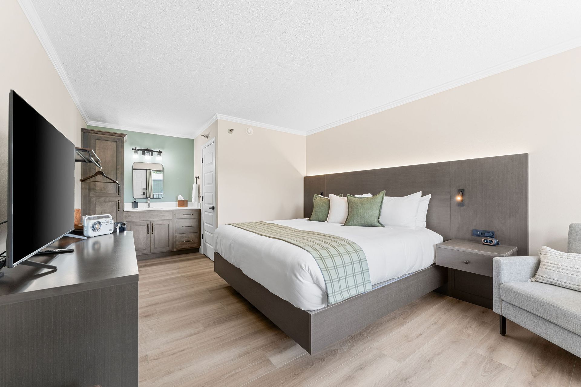 A modern hotel room with a king bed, a gray headboard, a large television, and an open-concept vanity area.