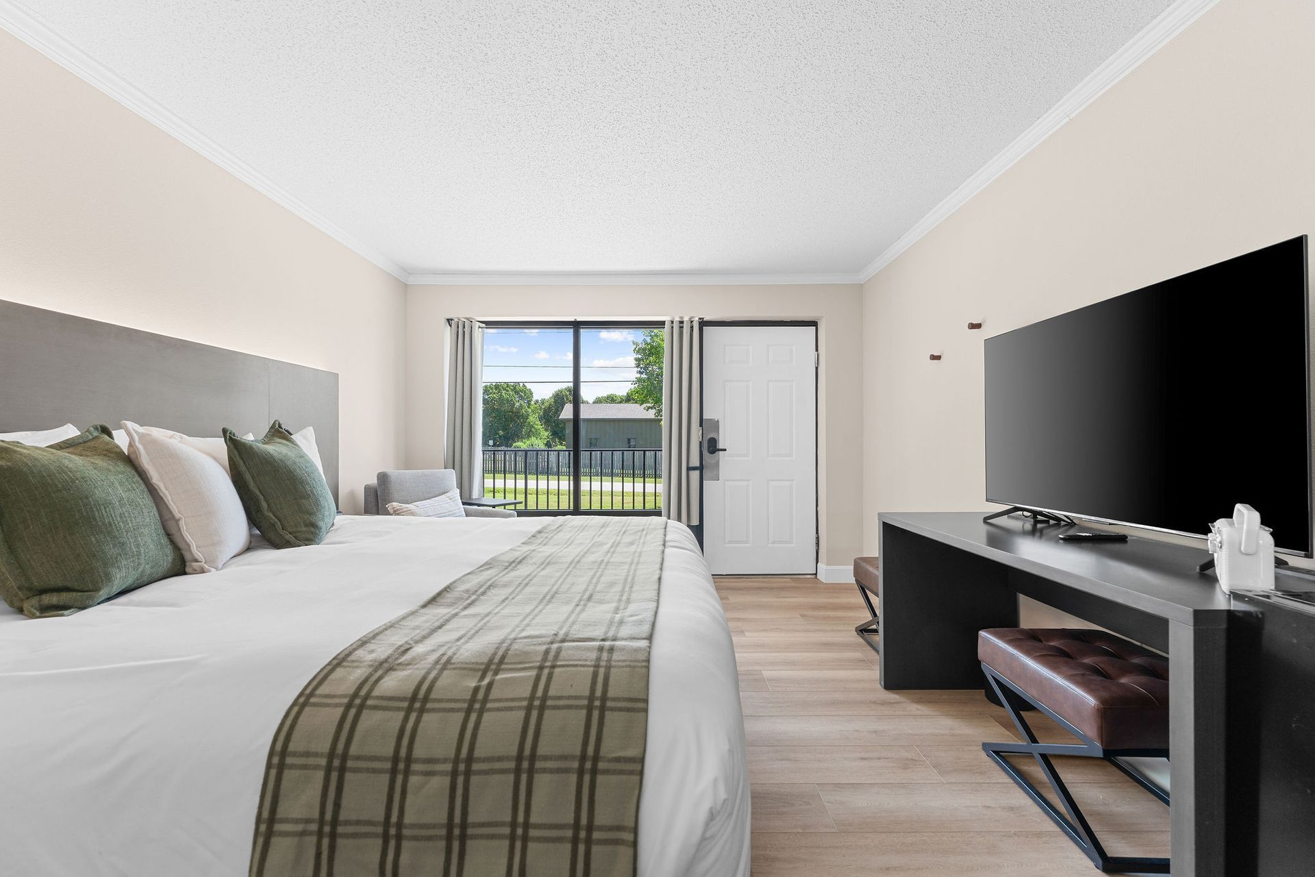 A modern hotel room with a large bed, olive green pillows, a plaid runner, a flat-screen TV, and a view of a balcony.