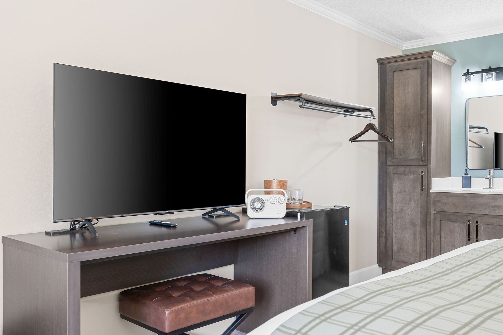 A modern hotel room view with a television on a dark desk, a brown stool, a hanging rack, a tall cabinet, and a vanity.