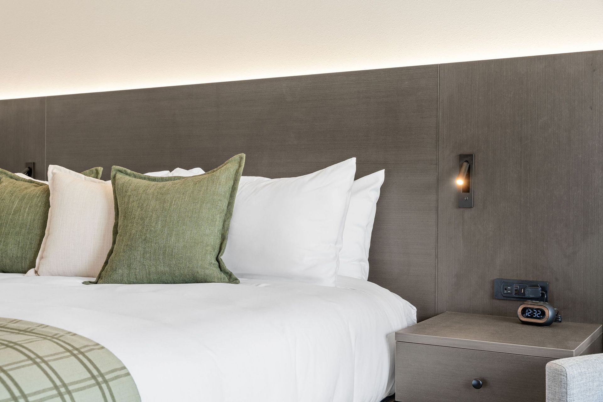A hotel bed with crisp white linens, green accent pillows, a plaid throw, and a modern wooden headboard with a reading light.