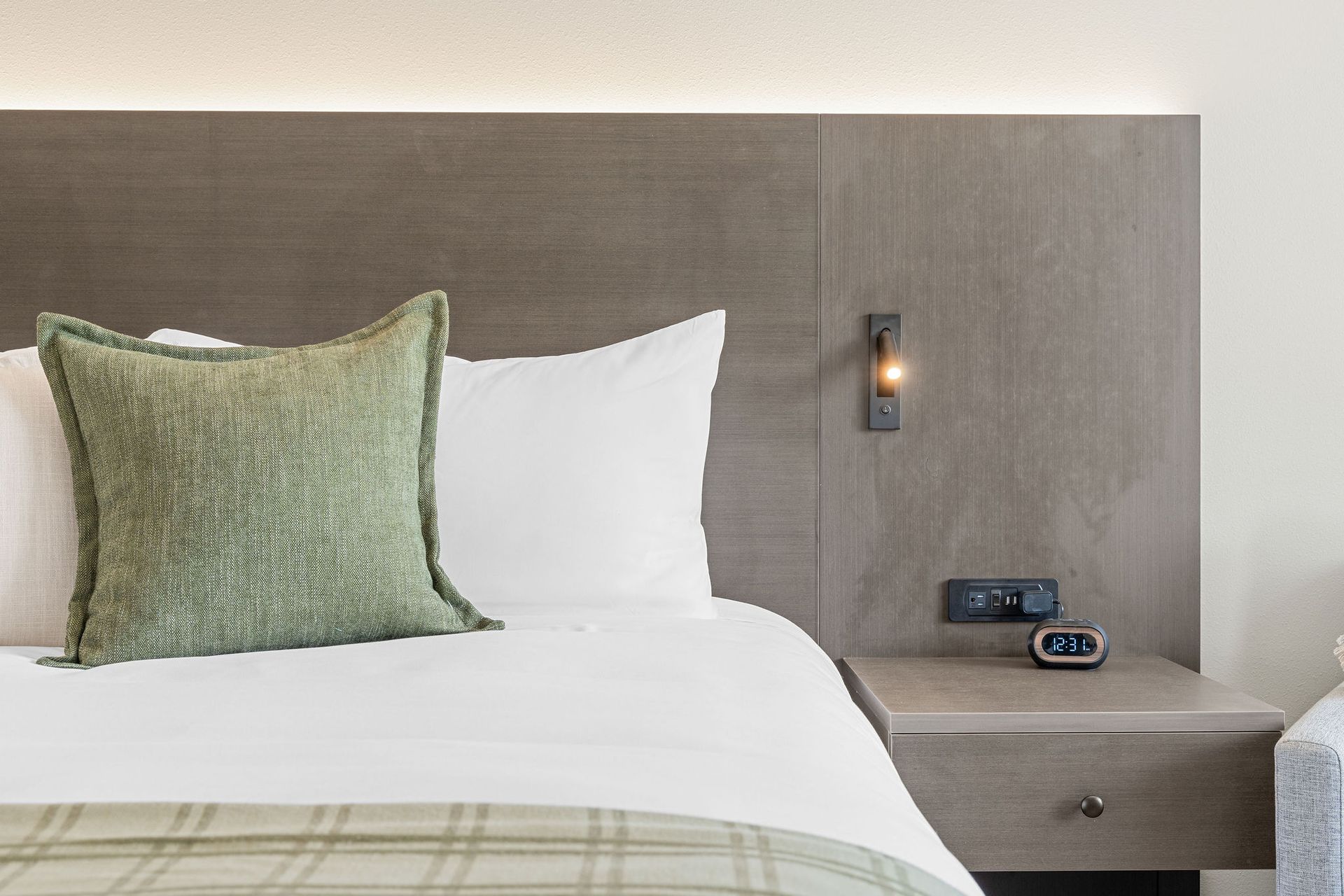 A hotel bed with white linens, a textured green accent pillow, wooden headboard, wall lamp, and a bedside clock.