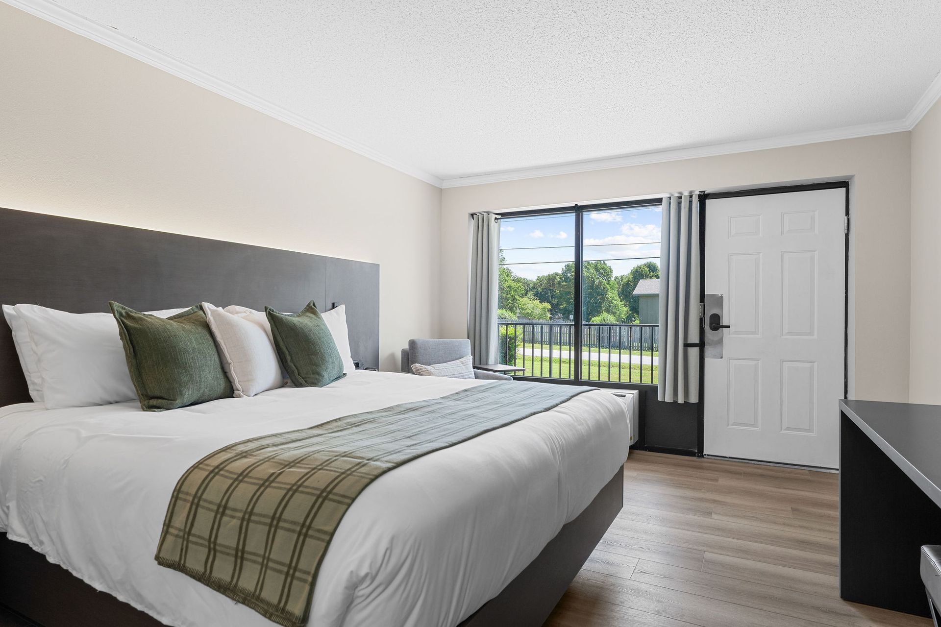 A modern hotel room with a large bed, light-colored walls, wood flooring, and a view of trees through a window and door.