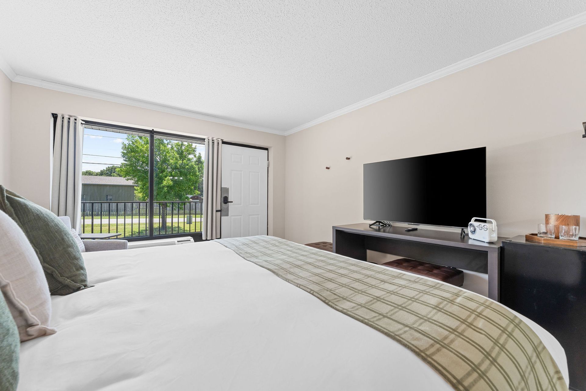 A brightly lit hotel room with a large bed in the foreground, facing a flat-screen TV on a wooden stand by a patio door.
