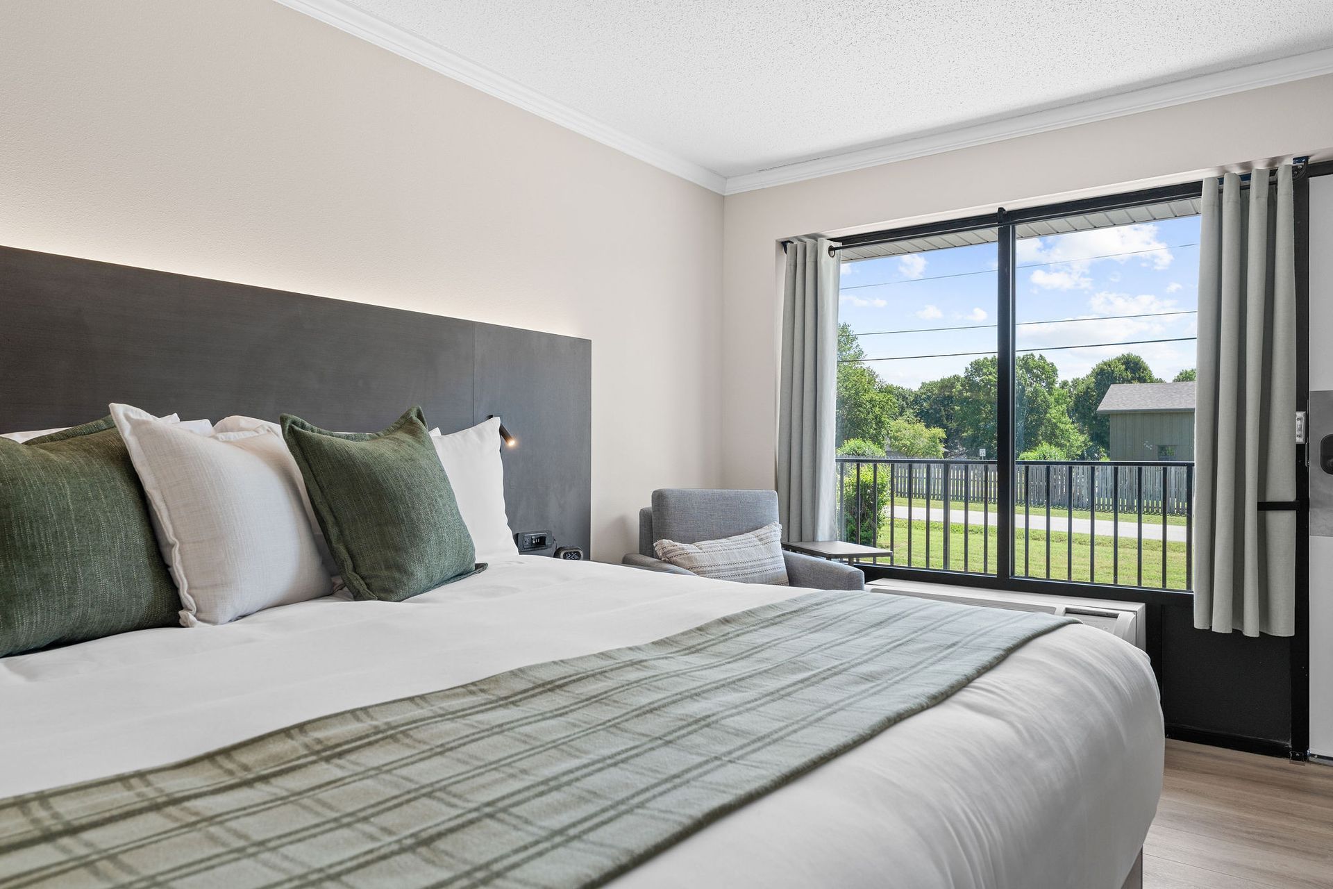 A neatly made hotel bed with white linens and sage green decorative pillows, next to a large window overlooking trees.