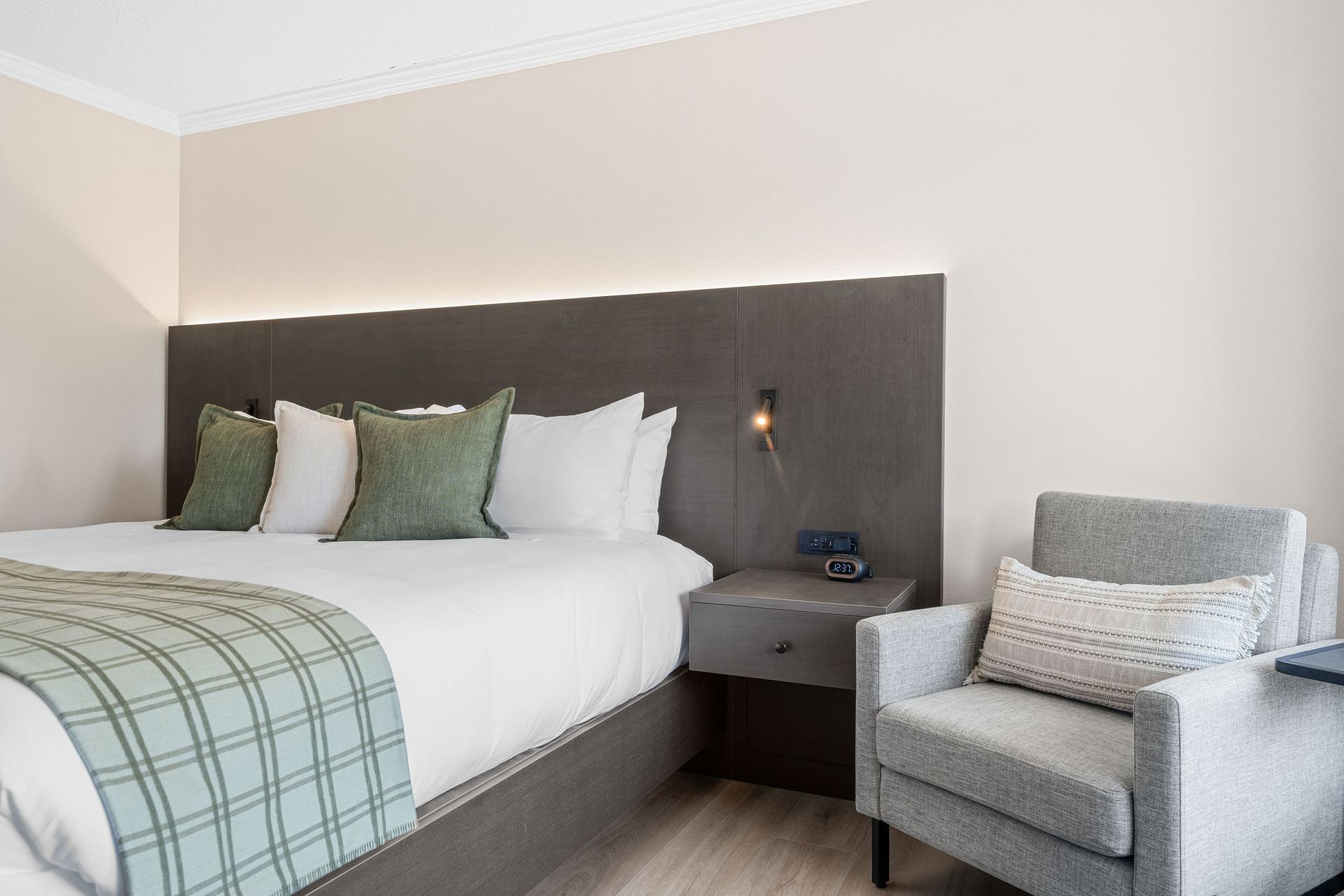 A hotel room featuring a bed with white bedding, green accent pillows and a throw, next to a gray armchair.