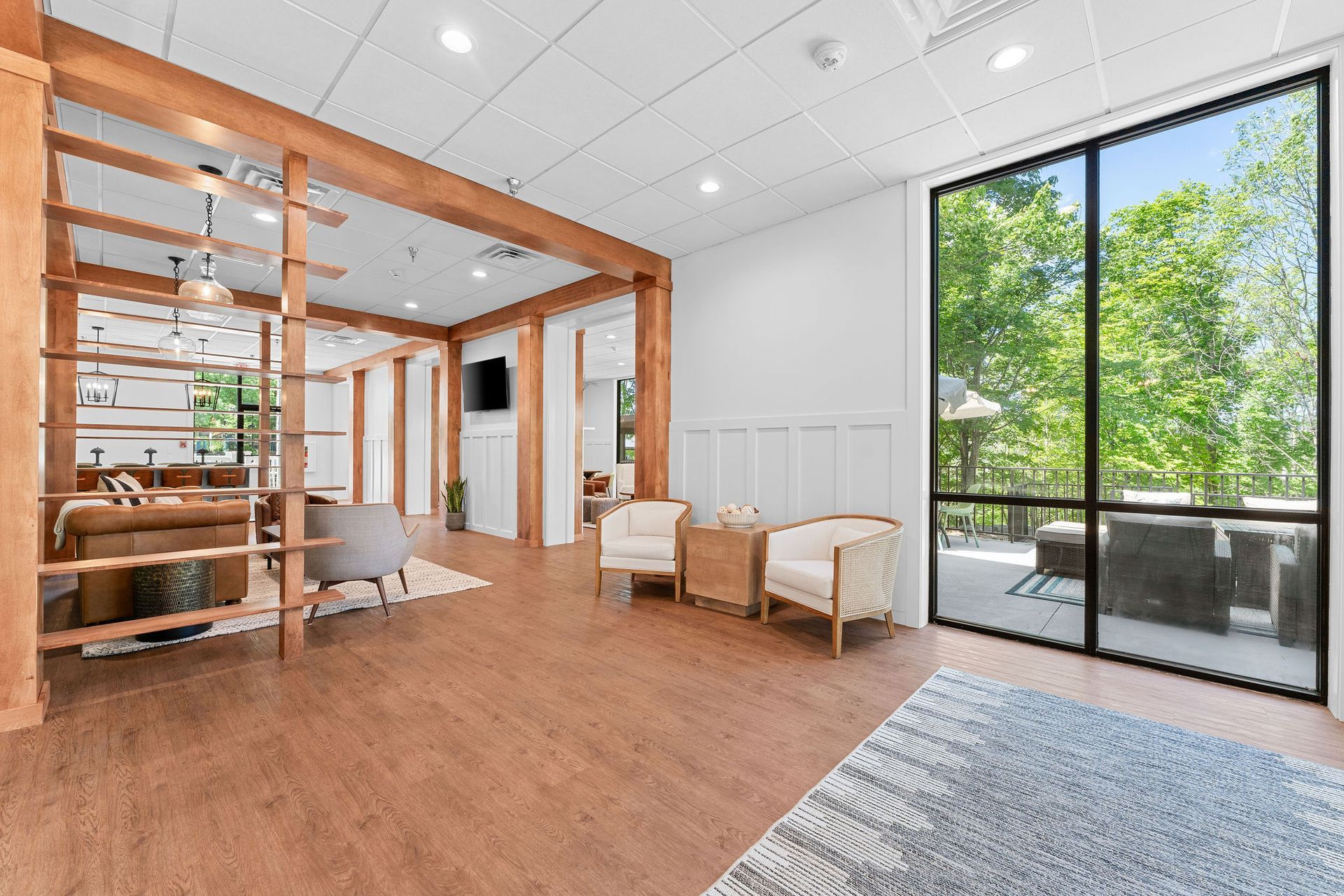 A modern open-plan lounge area with wooden room dividers, cream-colored armchairs, and a view of trees through a window.