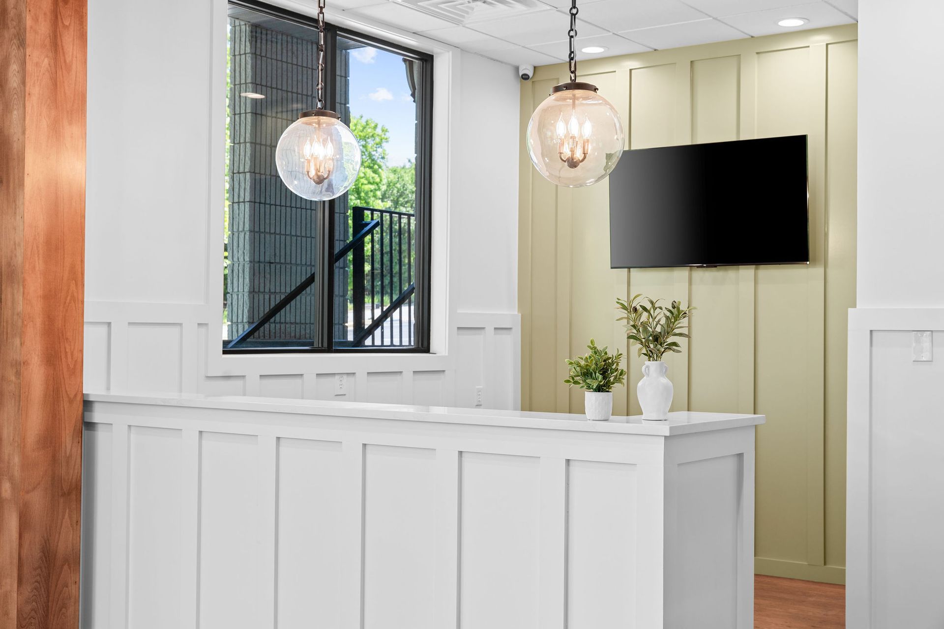 A bright, modern reception desk with a white panel design, globe pendant lights, two small plants, and a wall-mounted TV.