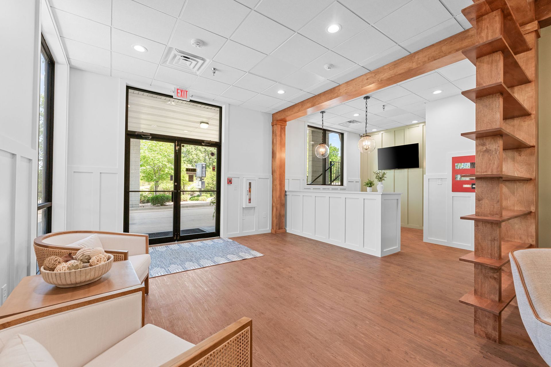 A brightly lit lobby with a white reception desk, wood-look flooring, glass entry doors, and a rustic wooden shelf unit.