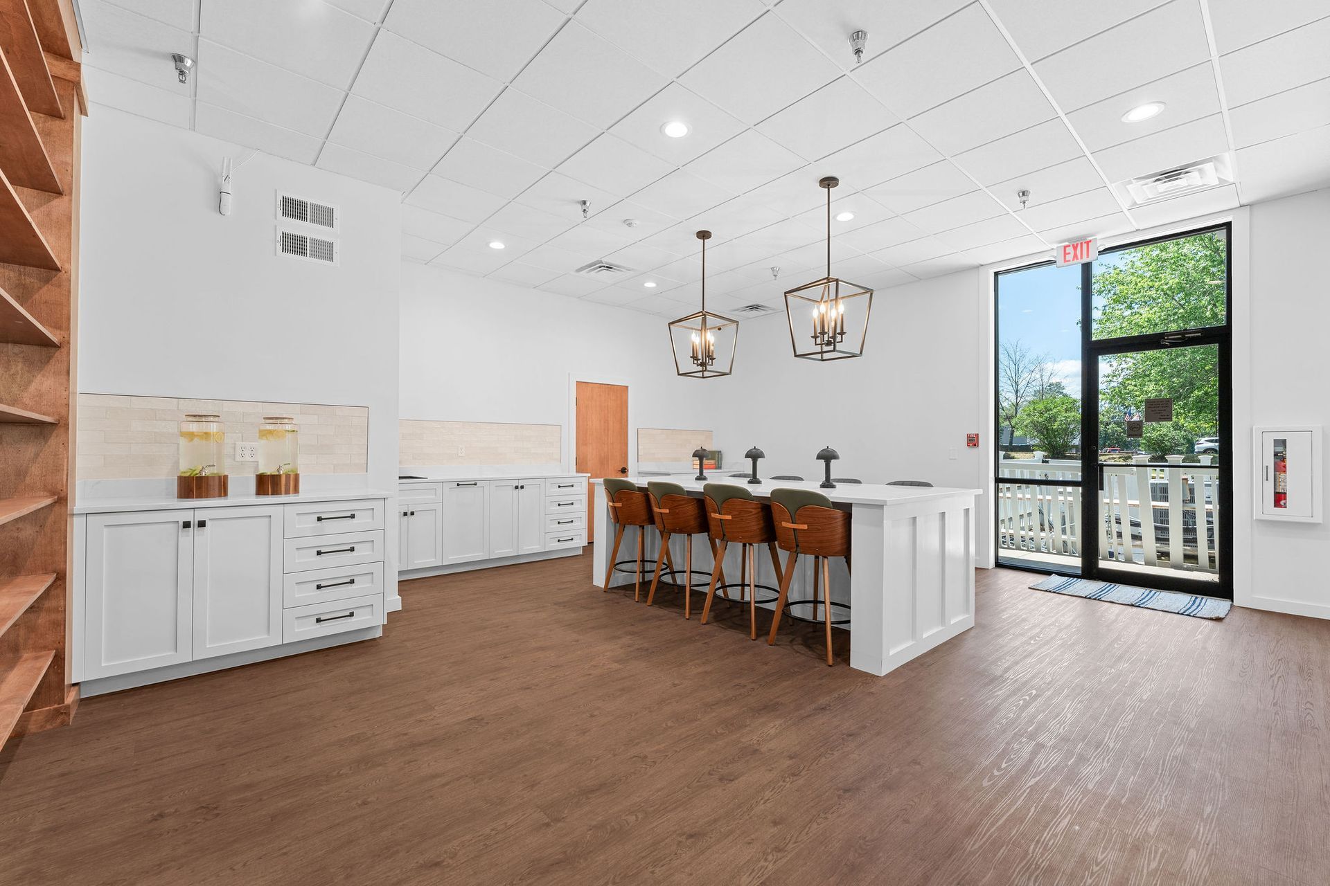 A bright kitchen space with a central island and four bar stools, wood-style flooring, and a glass door leading outside.