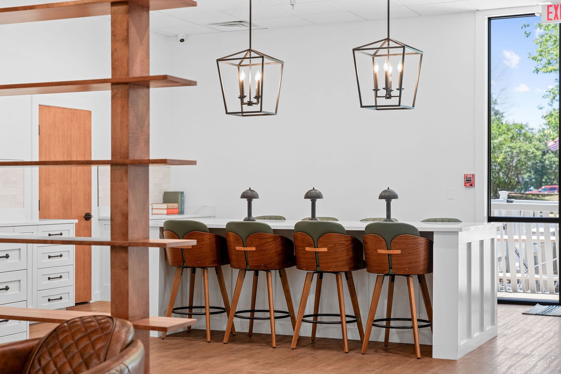 A modern office break area with a white bar counter, four brown stools, wooden shelving, and two pendant lanterns.