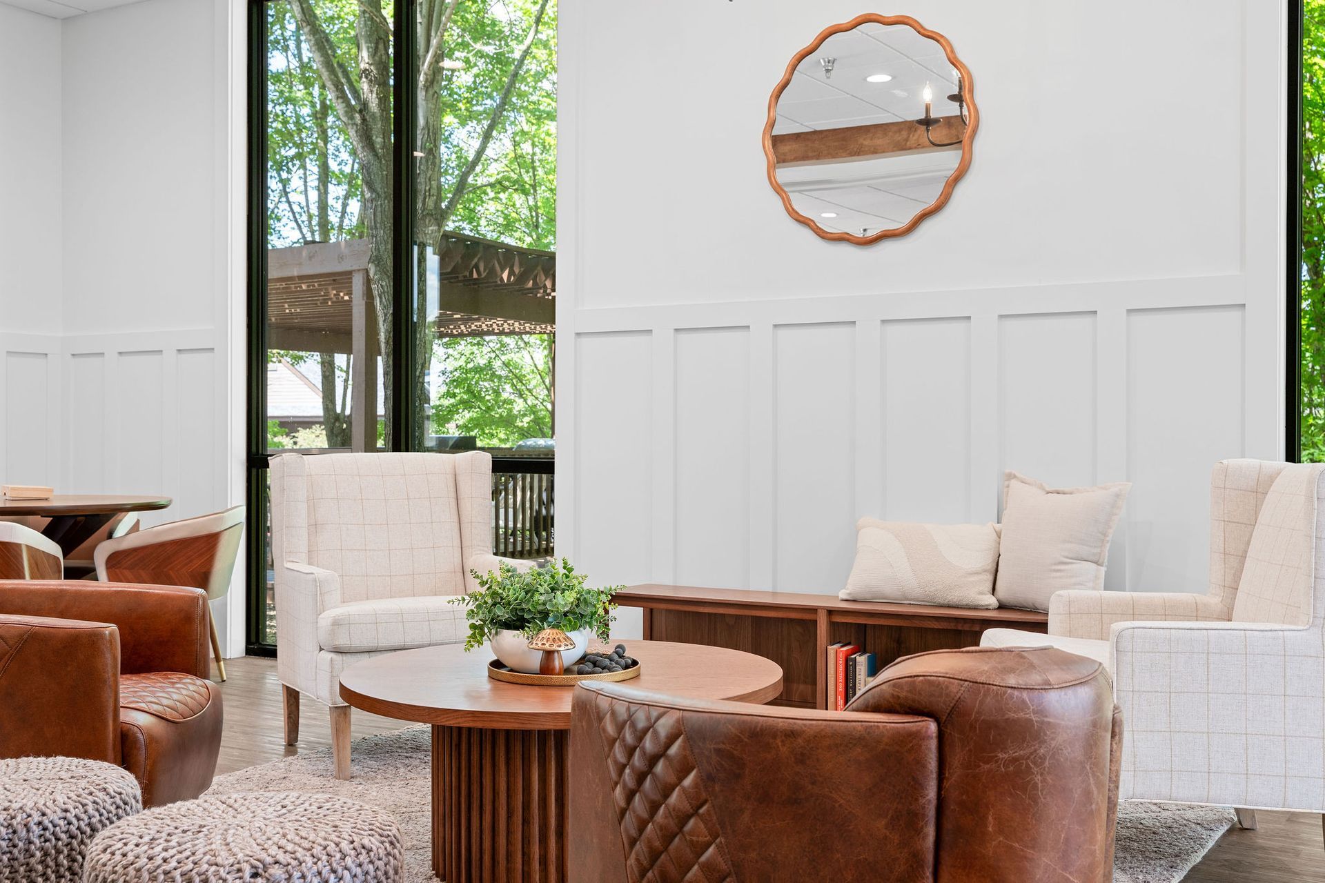 A lounge area features white chairs, brown leather armchairs, a circular wooden coffee table, and a round wall mirror.