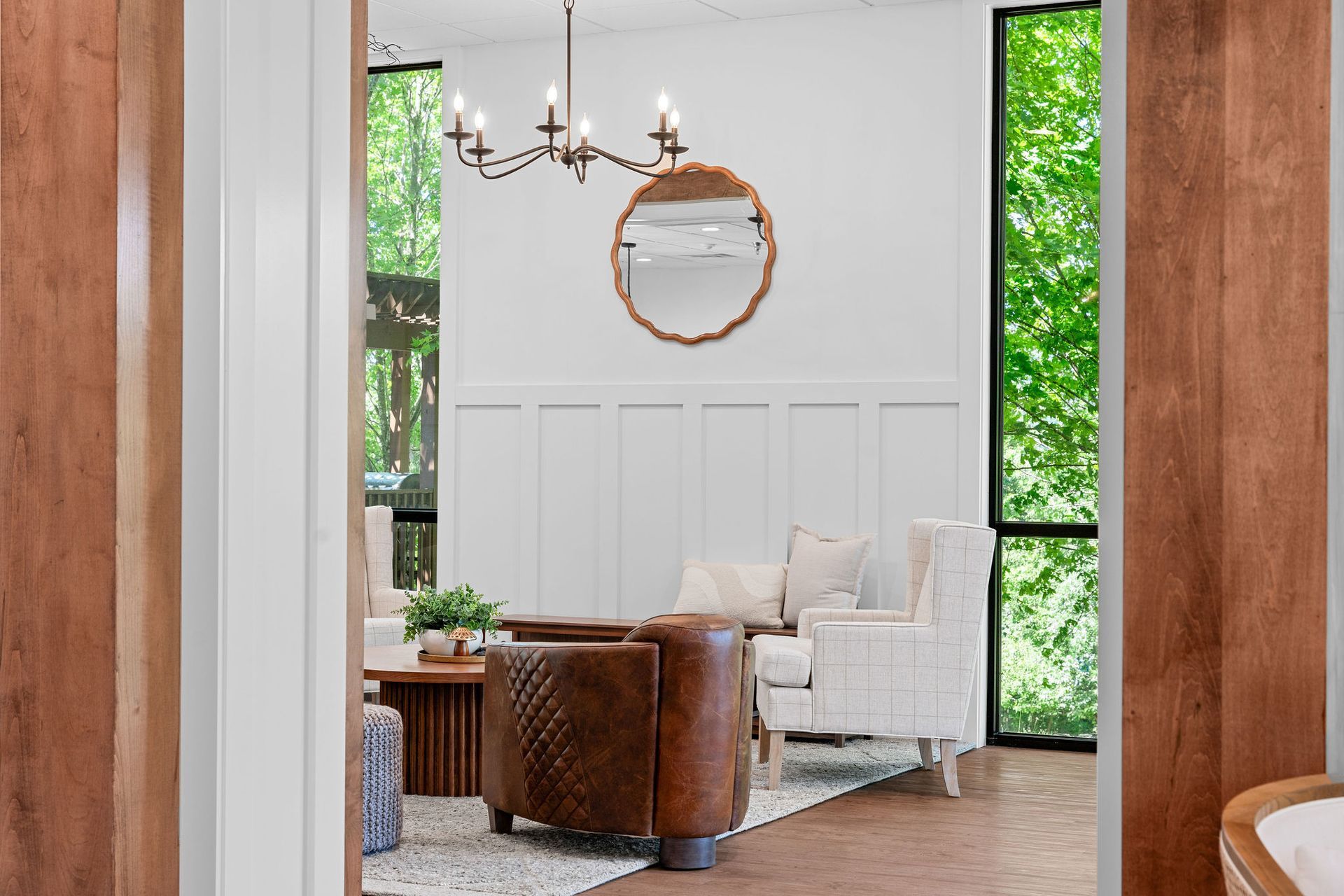 A bright, modern living space framed by wooden columns, featuring a brown leather chair, white sofa, and a round mirror.