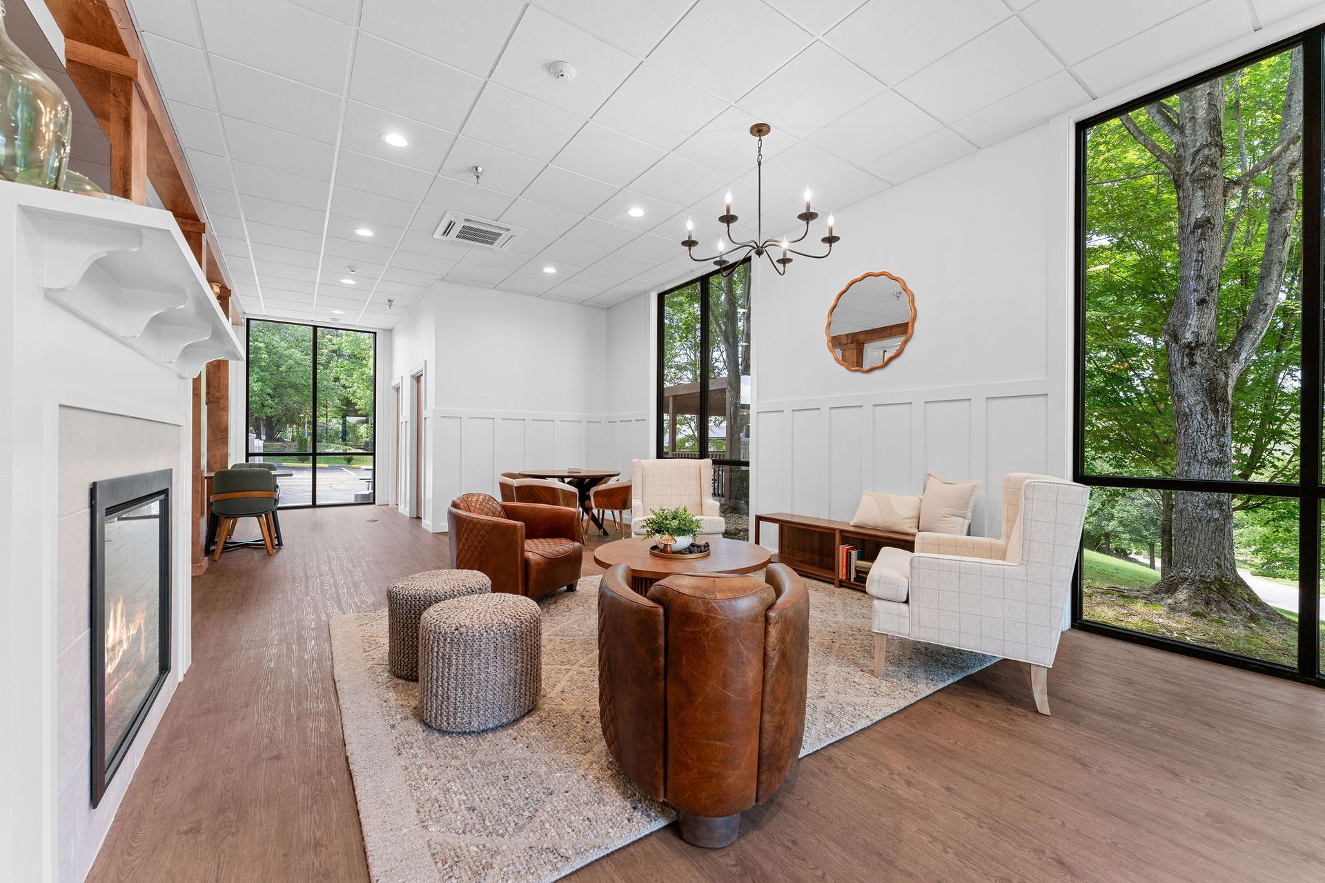 A spacious, bright lounge with a fireplace, comfortable seating, large windows overlooking trees, and a modern chandelier.