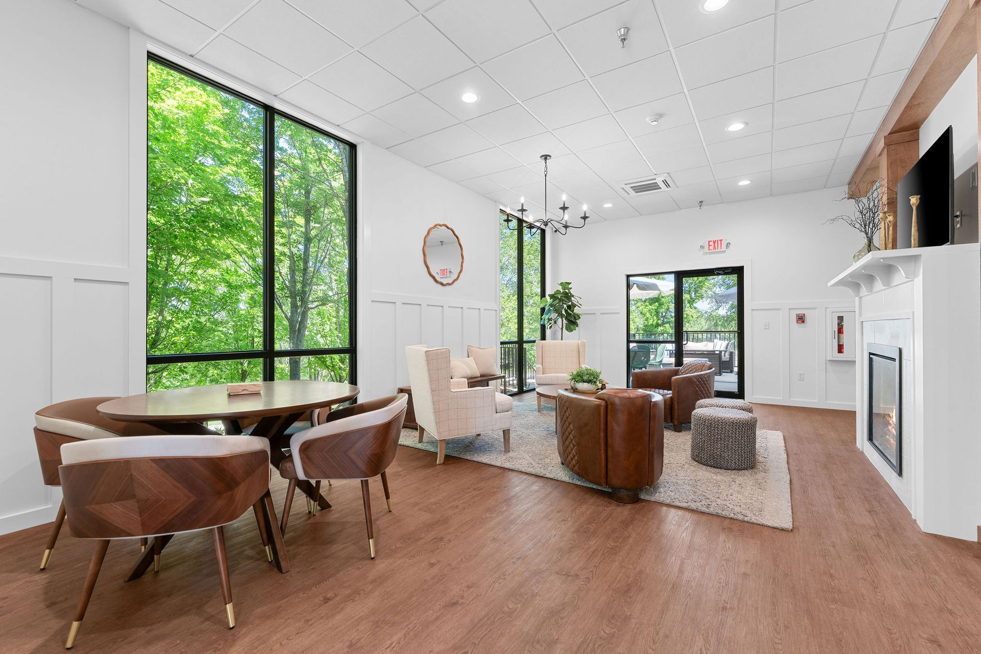 A modern lobby area with wood flooring, white walls, a dining table, lounge seating around a fireplace, and large windows.
