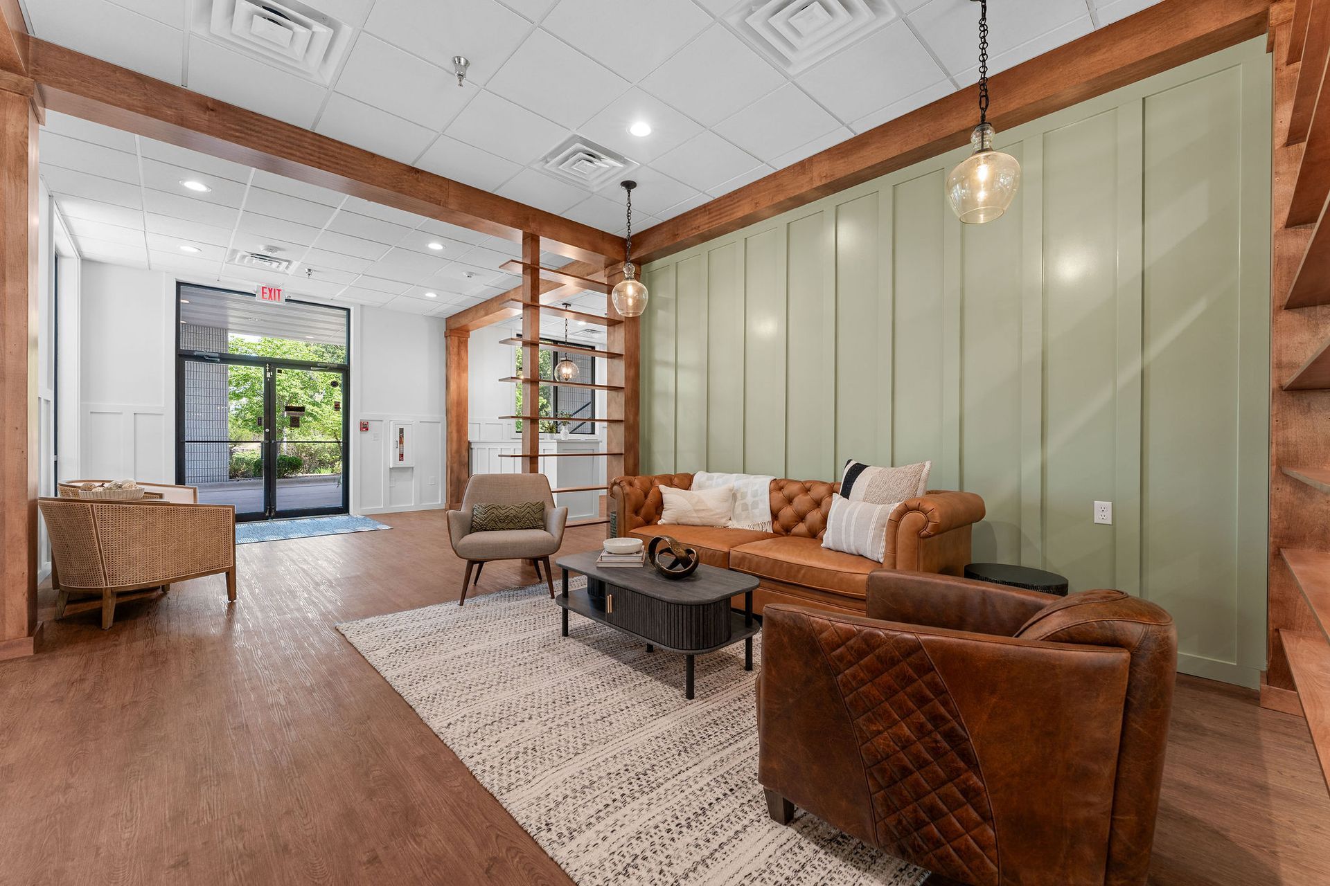 A modern lobby with a leather sofa, two armchairs, a patterned rug, and a light green paneled wall near a glass entrance.