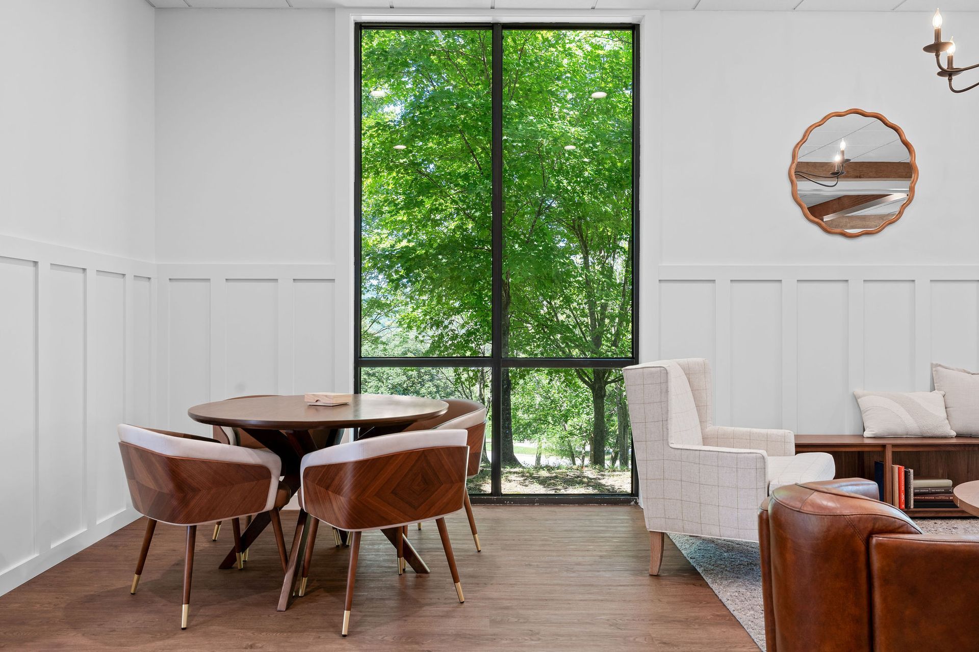 A brightly lit, modern room features white wainscoting, a dining set, neutral armchairs, and a large window view of trees.