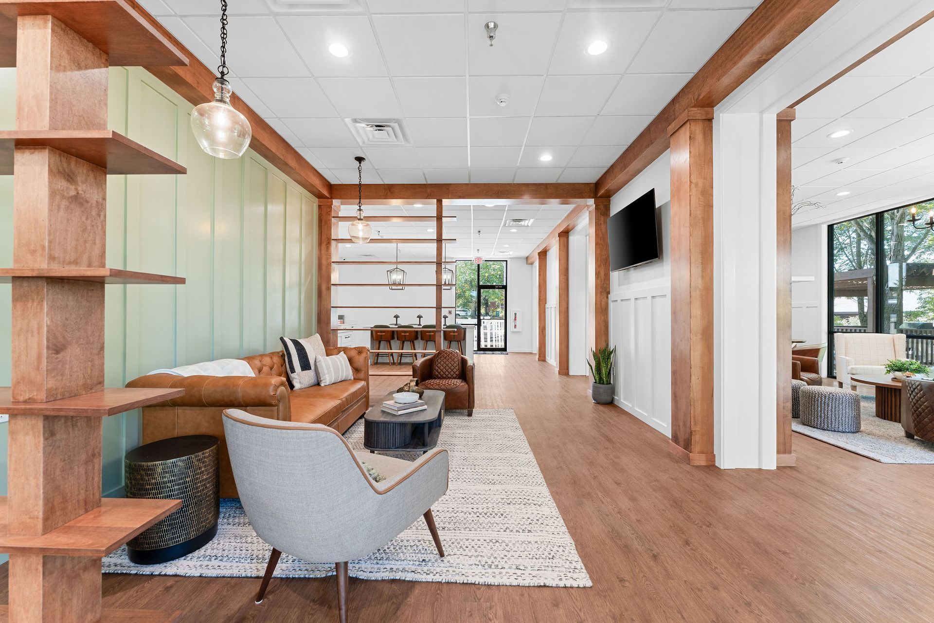 A modern, open-concept living area with a tan leather sofa, grey armchair, wooden shelving, and light wood flooring.