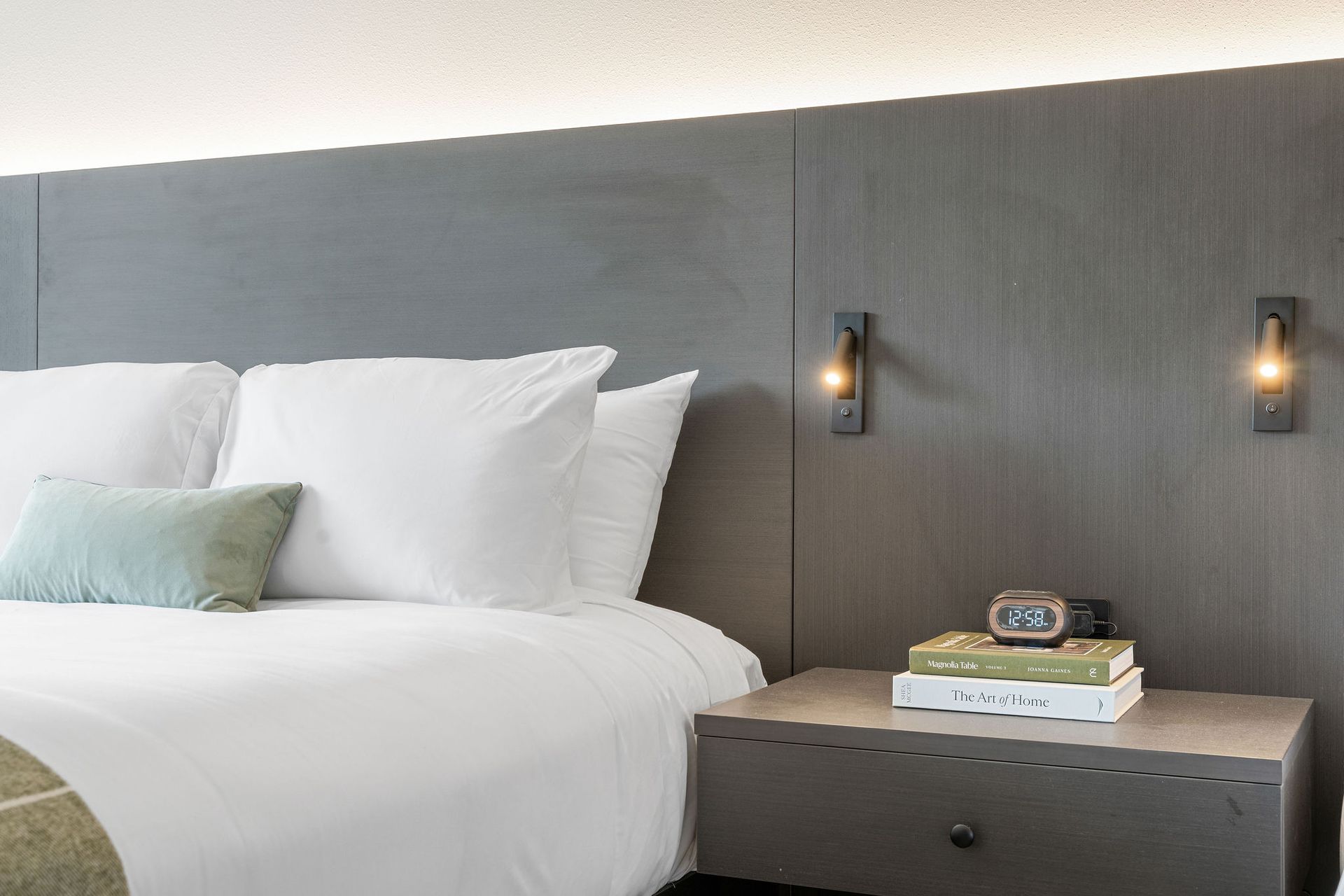 A modern hotel bed with white linens, pillows, a sage accent cushion, and a dark wood headboard with built-in wall lamps.