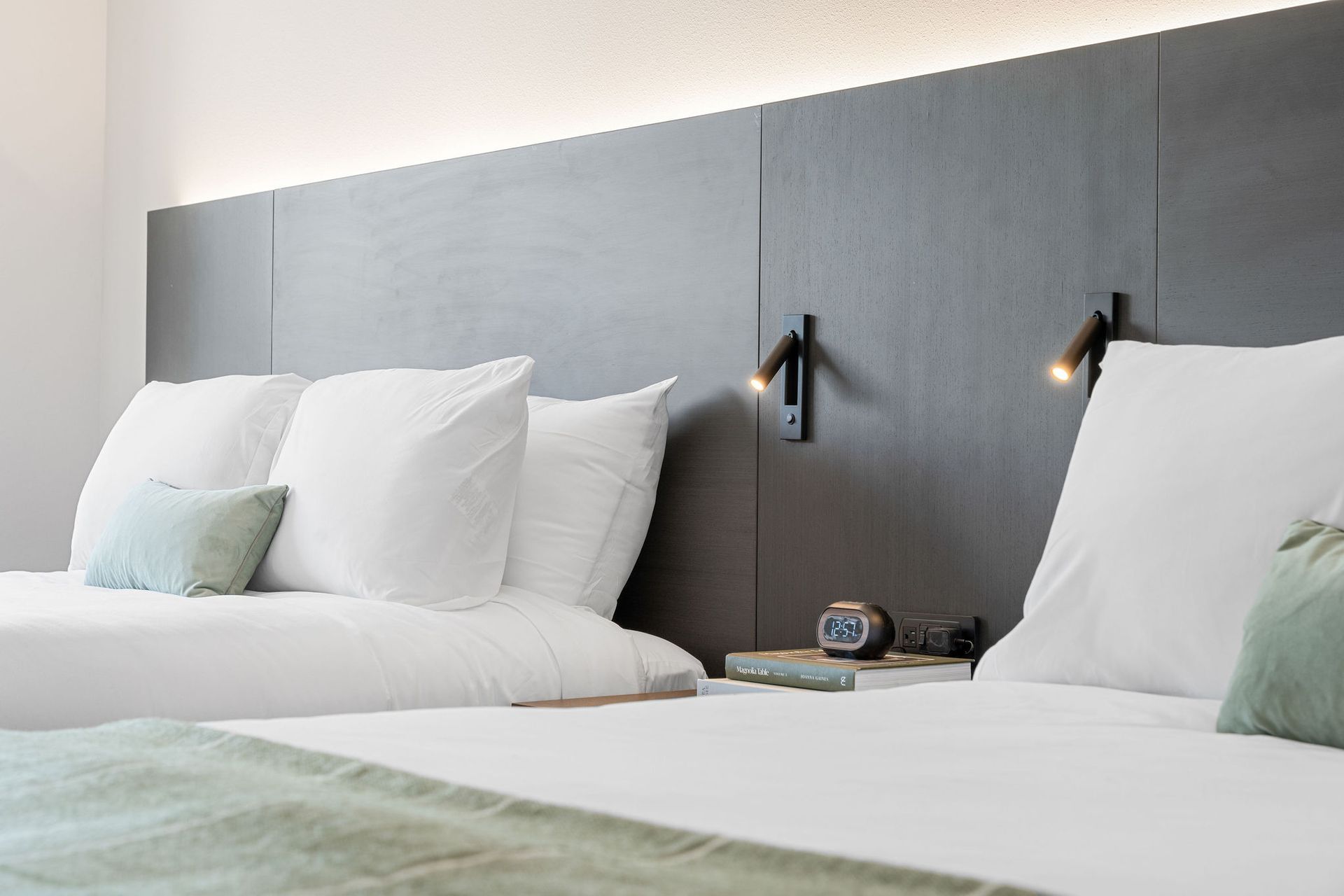 A modern hotel room with two white beds, a dark headboard, and minimalist reading lights.