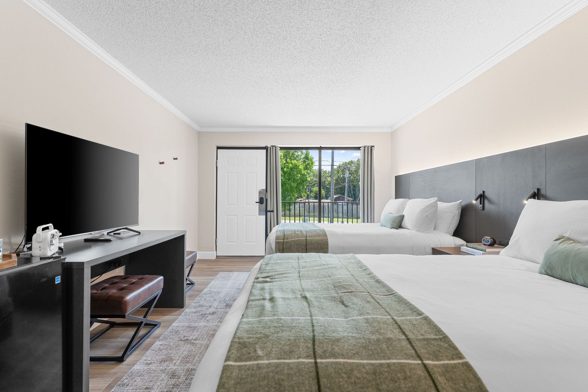 A bright, modern hotel room with two queen beds, a television on a long desk, and an exterior door leading to a balcony.