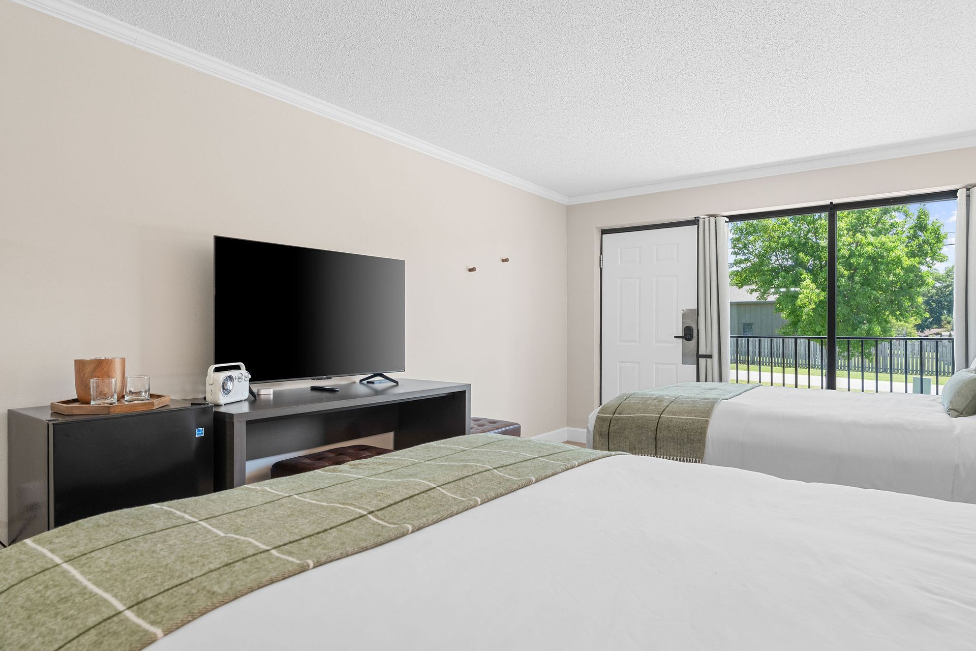 A bright hotel room featuring two beds, a black TV stand with a television, and a sliding glass door with a balcony view.
