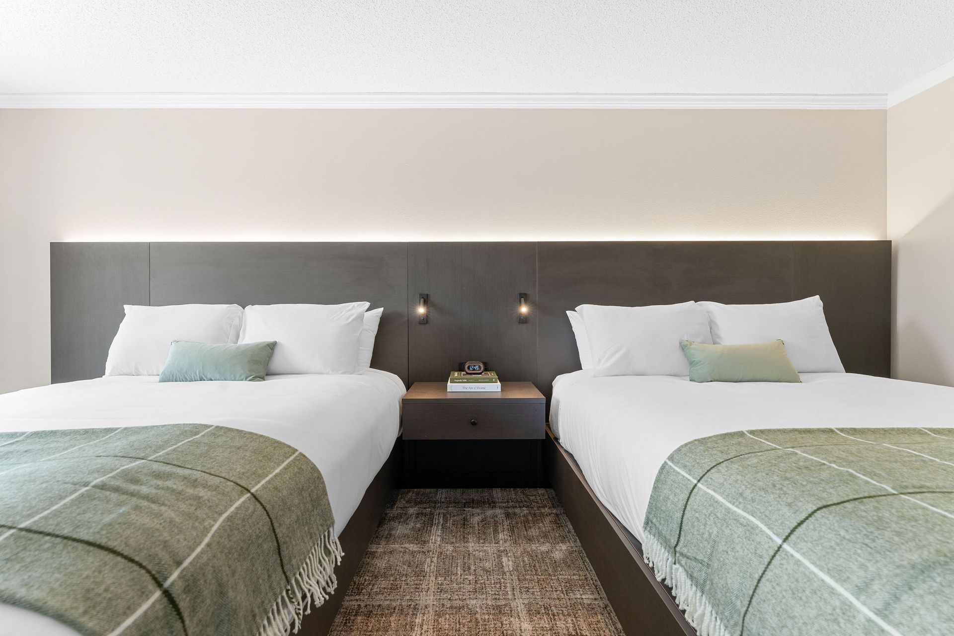 Two beds with white linens and green plaid throws, separated by a dark wood nightstand against a dark paneled headboard.