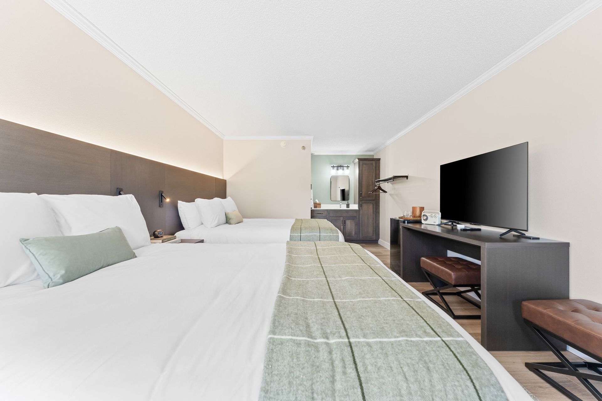 A hotel room with two queen beds featuring white linens, plaid runners, a flat-screen TV, and an adjacent bathroom area.