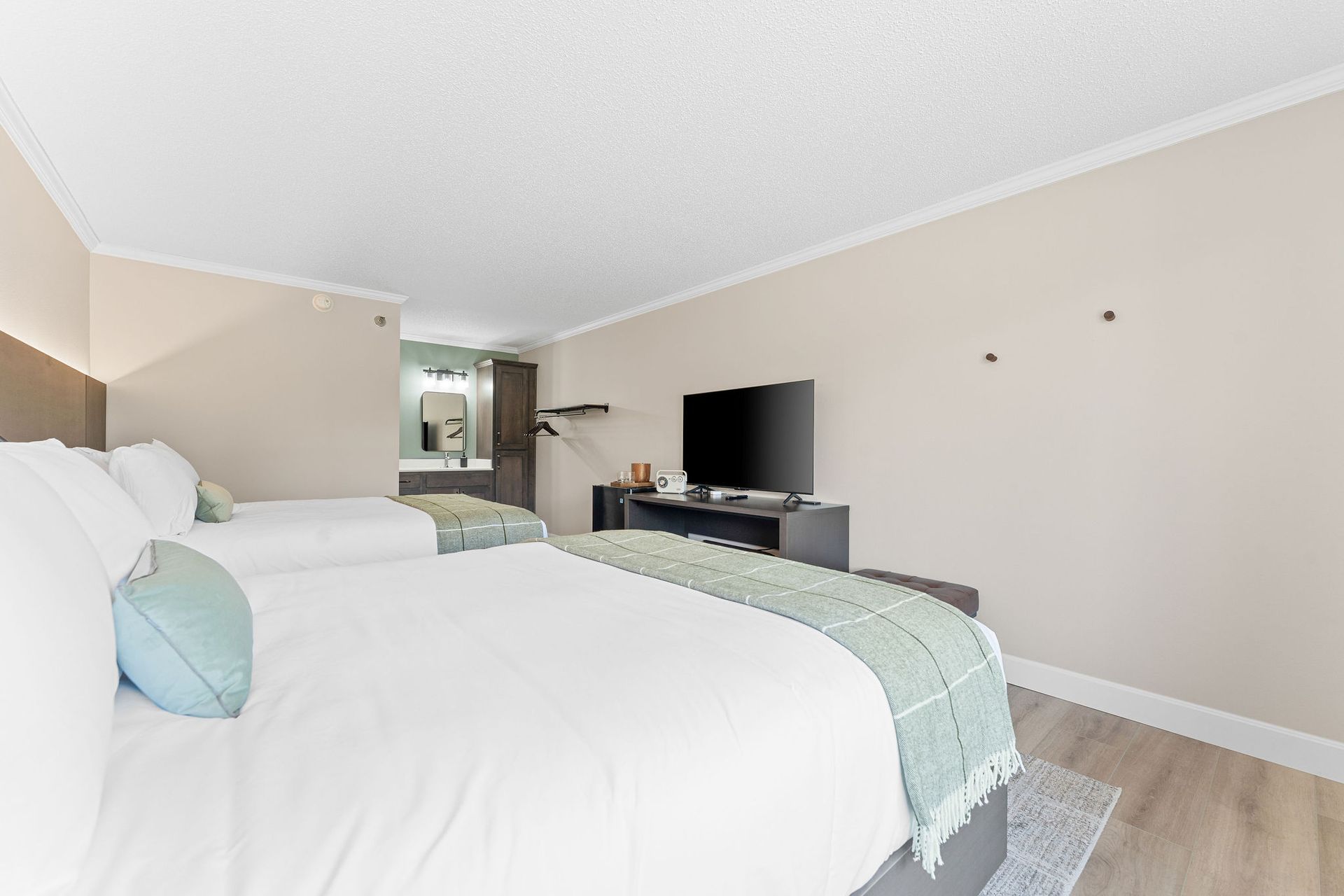 A hotel room with two beds, featuring white linens and light blue accents, a television on a stand, and a vanity area.